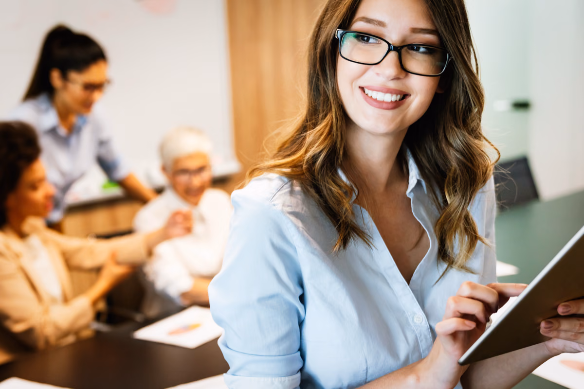 A woman holding a tablet in a meeting room, with colleagues working at the table behind her.