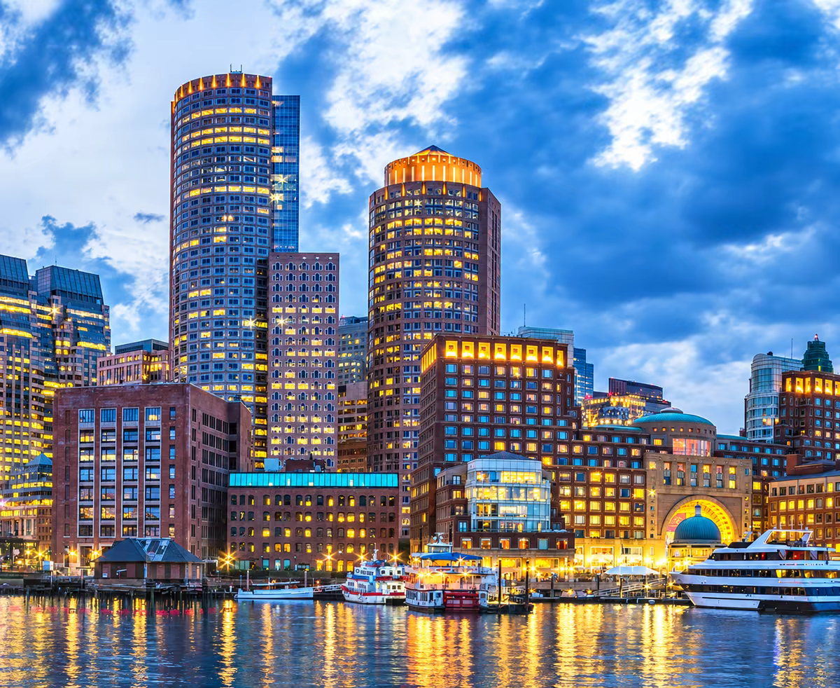 The Boston skyline at dusk, seen across the harbour with the lit towers of the financial district reflected in the water.