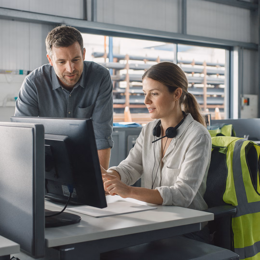 Two colleagues at a desk looking at the same monitor in a sales office, a headset around one of their necks.