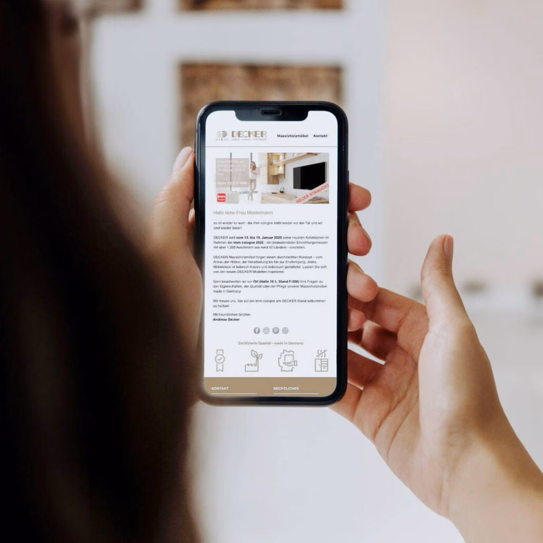Hands hold a phone showing a Decker e-mail newsletter that greets a sample recipient by name and invites her to the company's trade fair stand.
