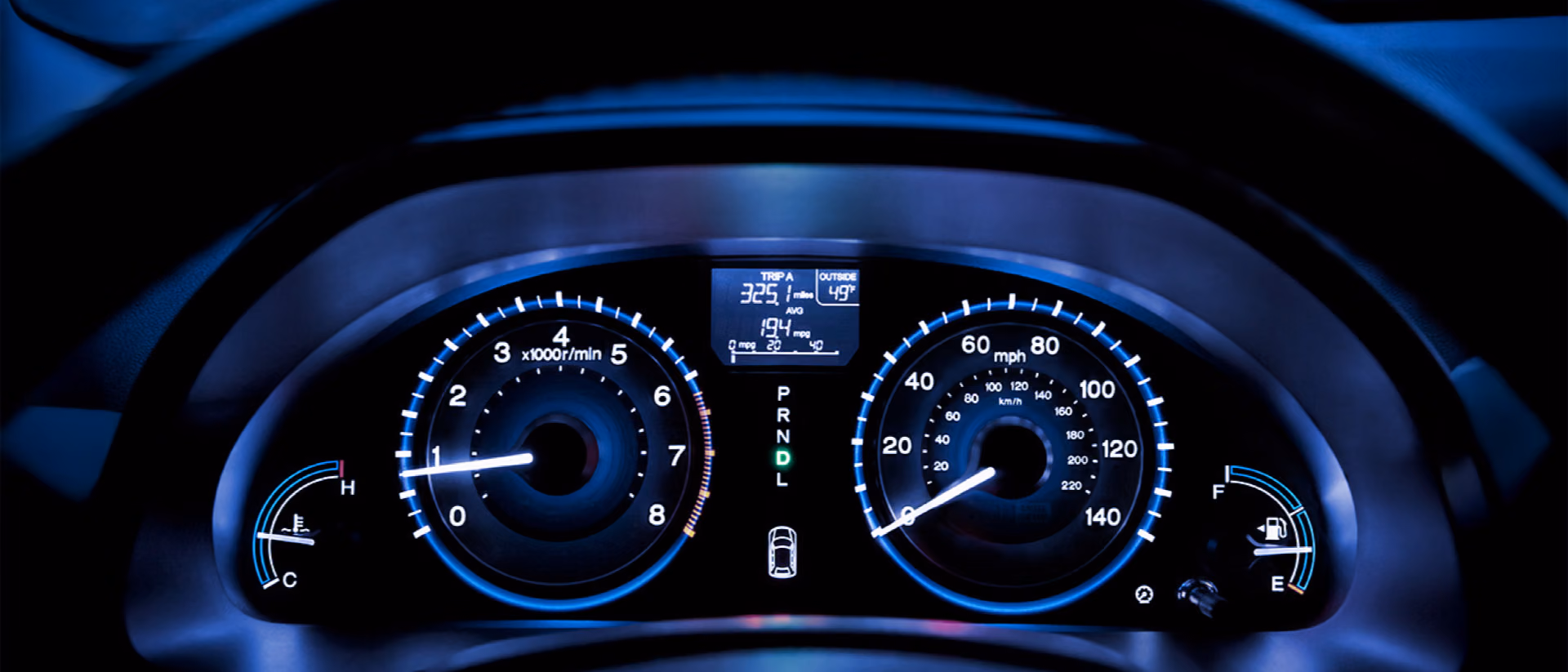 A car dashboard at night, tachometer and speedometer lit in blue either side of a small trip display.