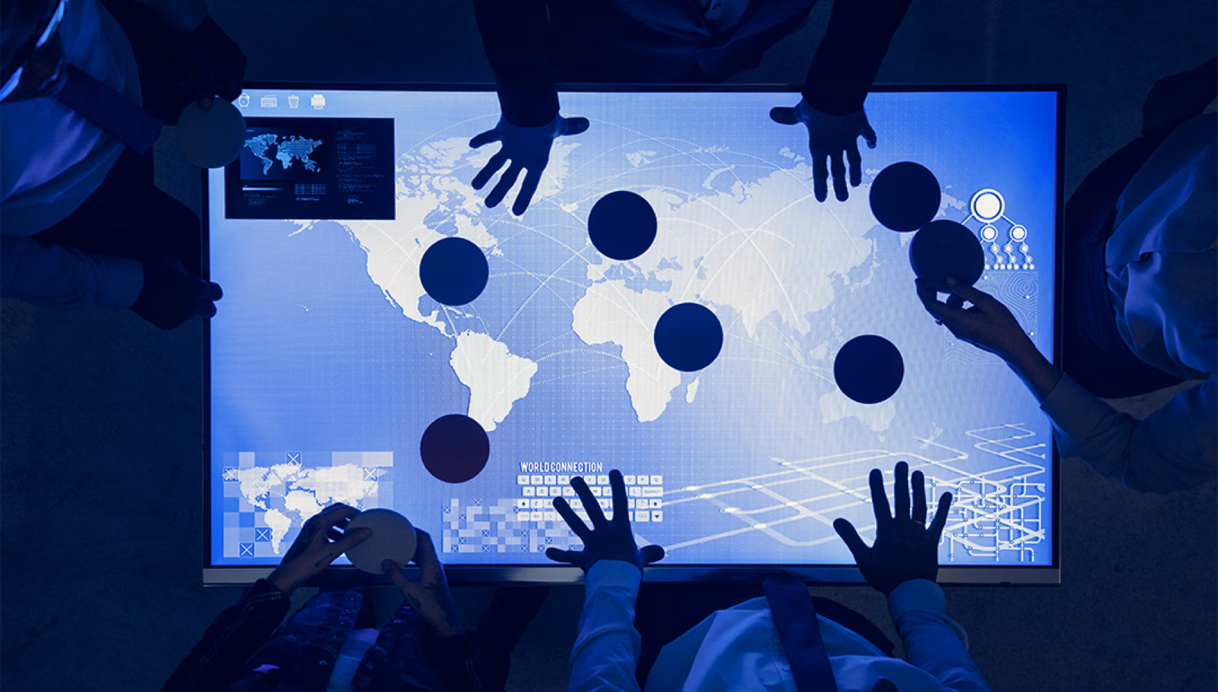 Seen from above, several people stand around a large lit touch table showing a world map and move dark circular tokens across it.