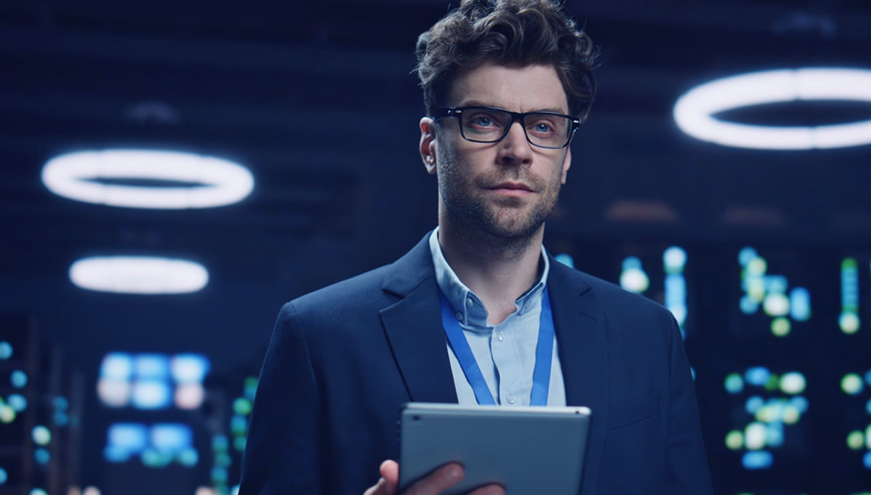 A man in a blazer and lanyard holds a tablet in a dark data centre, rows of status lights and ring lamps behind him.