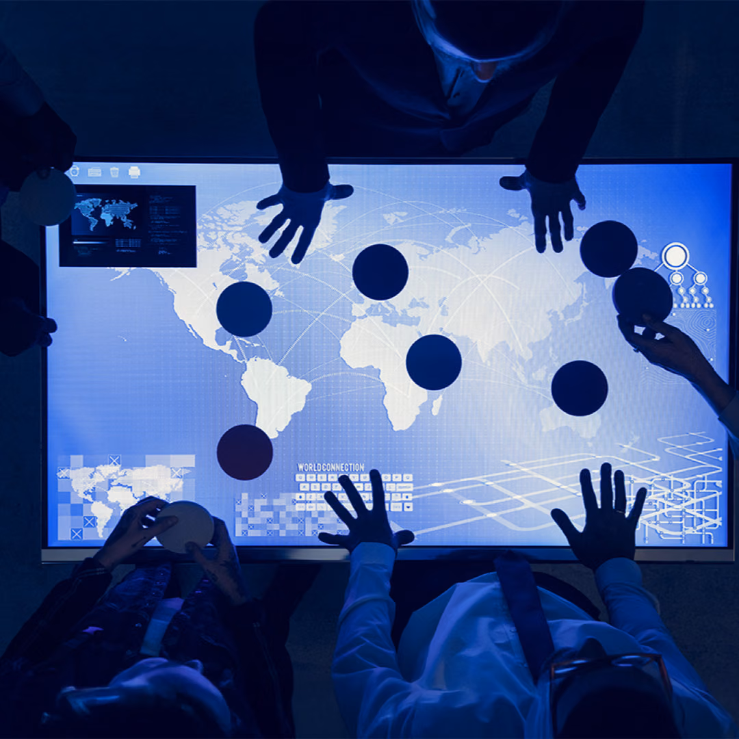 Seen from above, several people stand around a large lit touch table showing a world map and move dark circular tokens across it.
