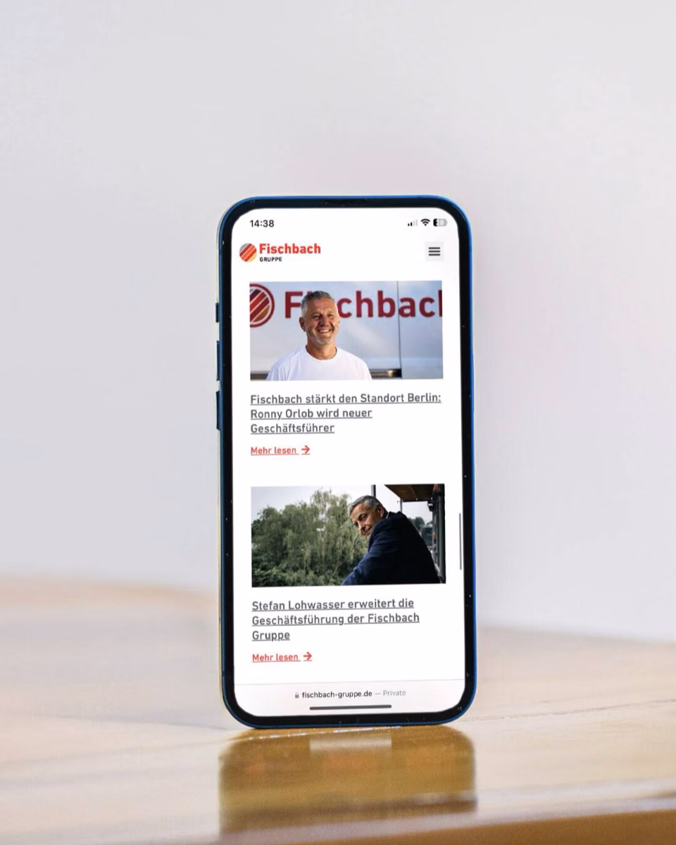 A phone standing on a wooden table shows the mobile Fischbach Gruppe site with two news items.