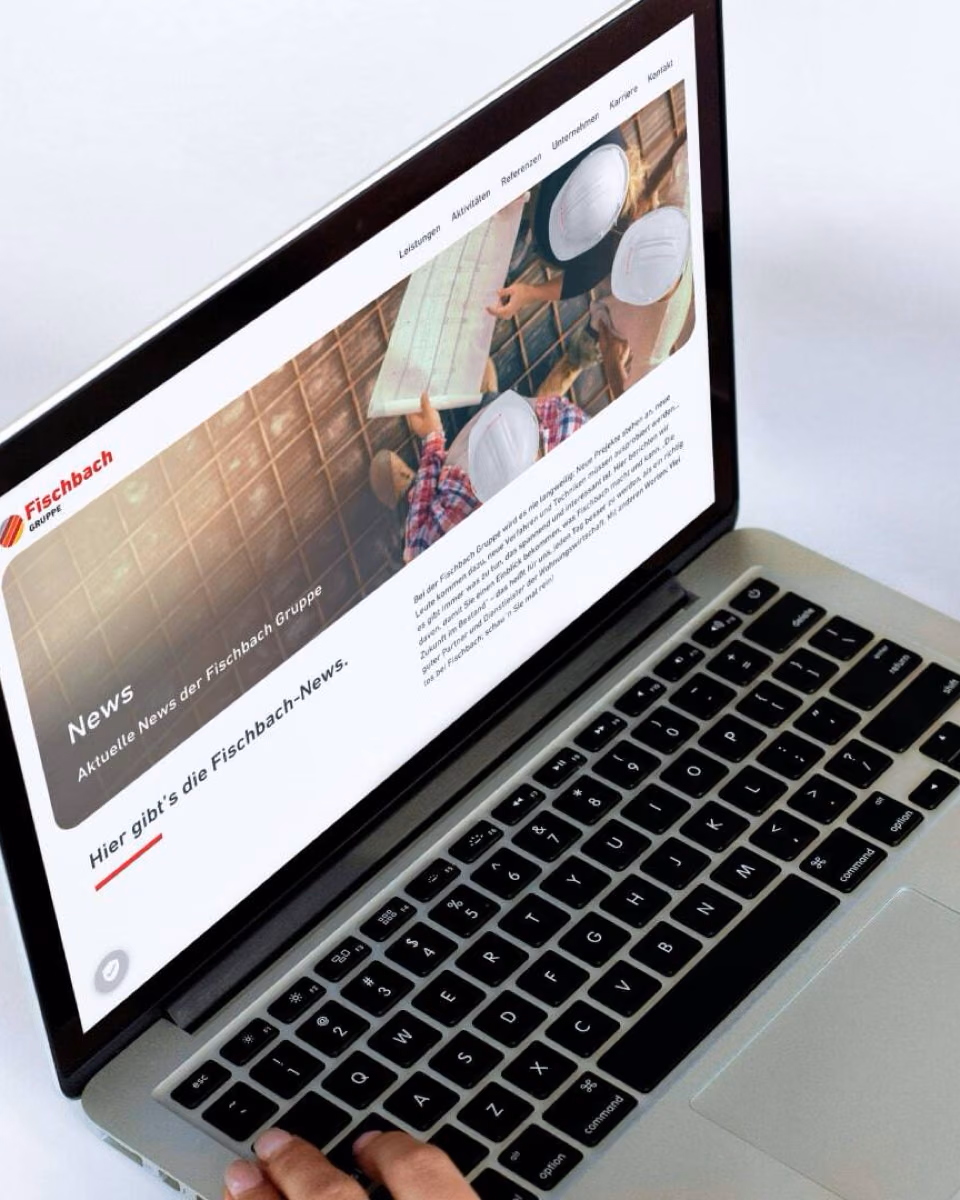 A hand types on a MacBook whose screen shows the Fischbach Gruppe news page.