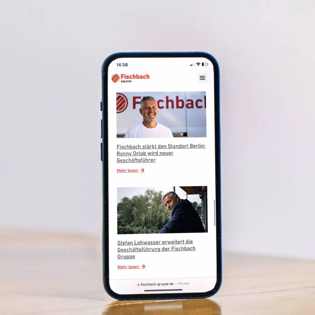 A phone standing on a wooden table shows the mobile Fischbach Gruppe site with two news items and their read-more links.