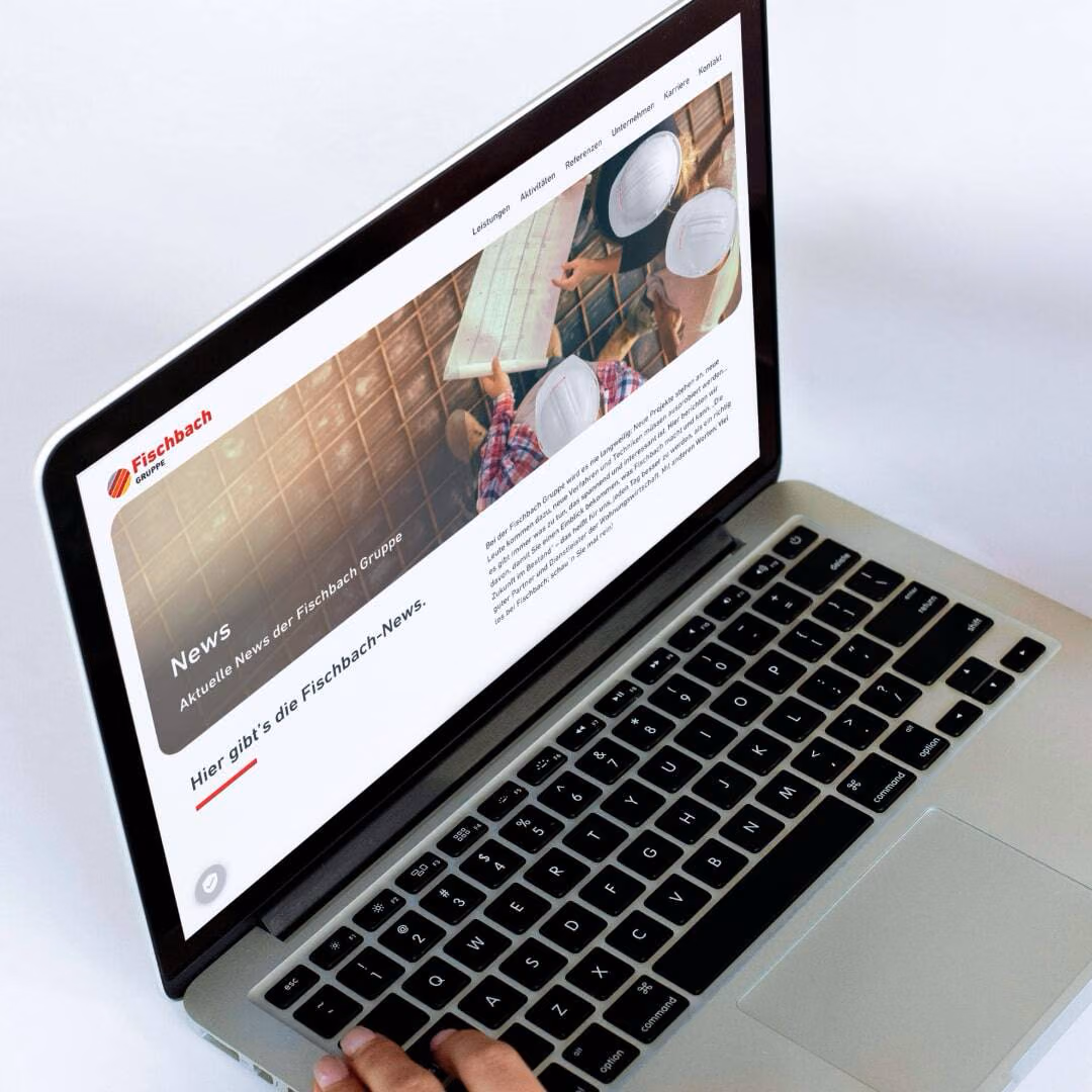 A hand types on a MacBook whose screen shows the Fischbach Gruppe news page.