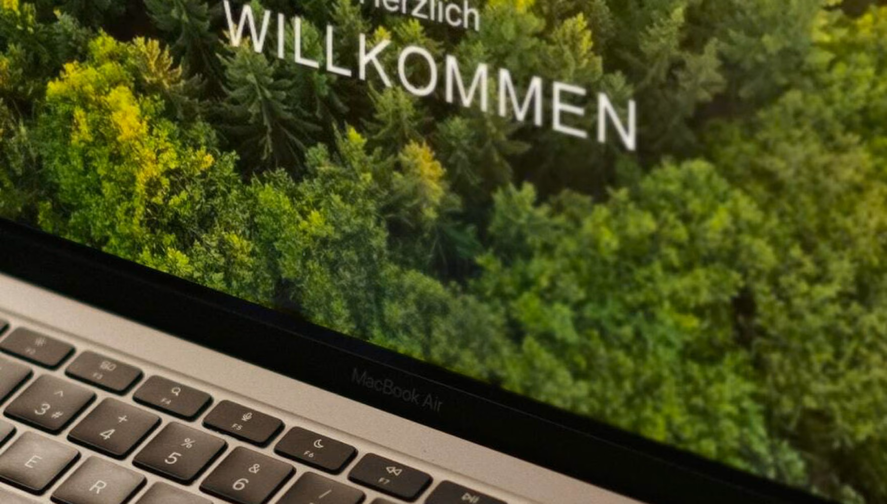 A close-up of a laptop screen greeting the reader with Herzlich Willkommen over an aerial photograph of a forest.