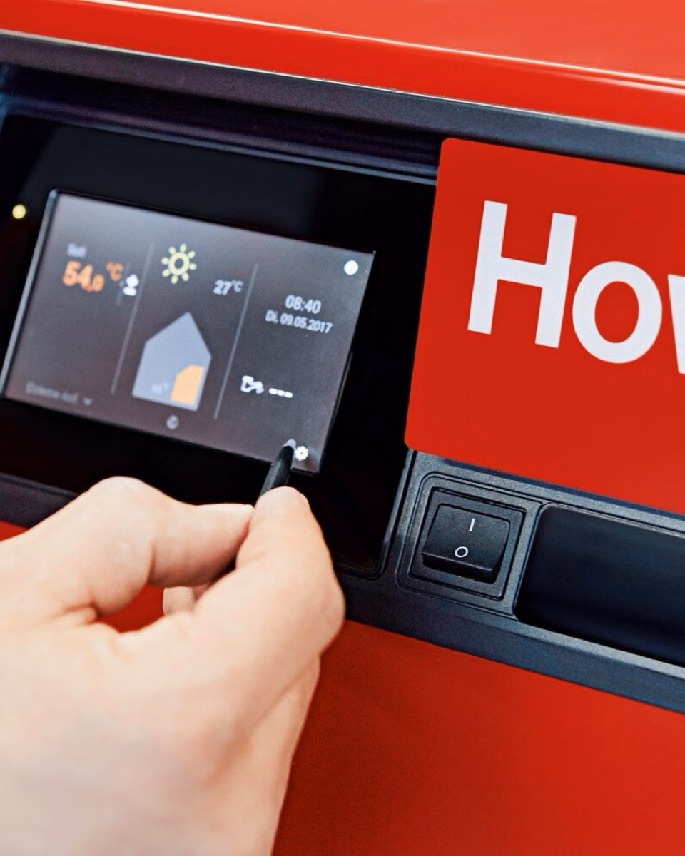 A hand reaches for the control display of a red Hoval boiler showing a flow temperature of 54 degrees.