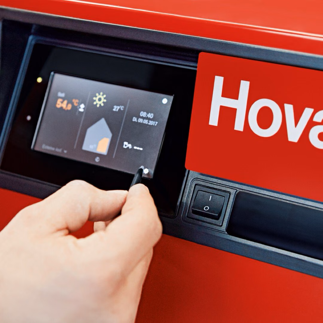 A hand reaches for the control display of a red Hoval boiler, which shows a flow temperature of 54 degrees beside a house symbol.