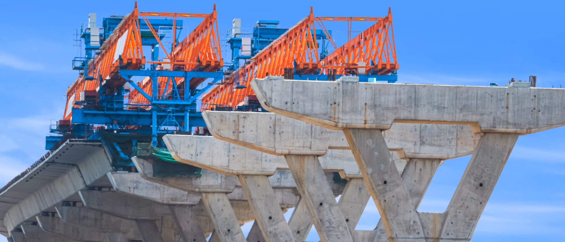 A bridge deck under construction, an orange launching gantry standing on a row of splayed concrete pier caps against a clear blue sky.