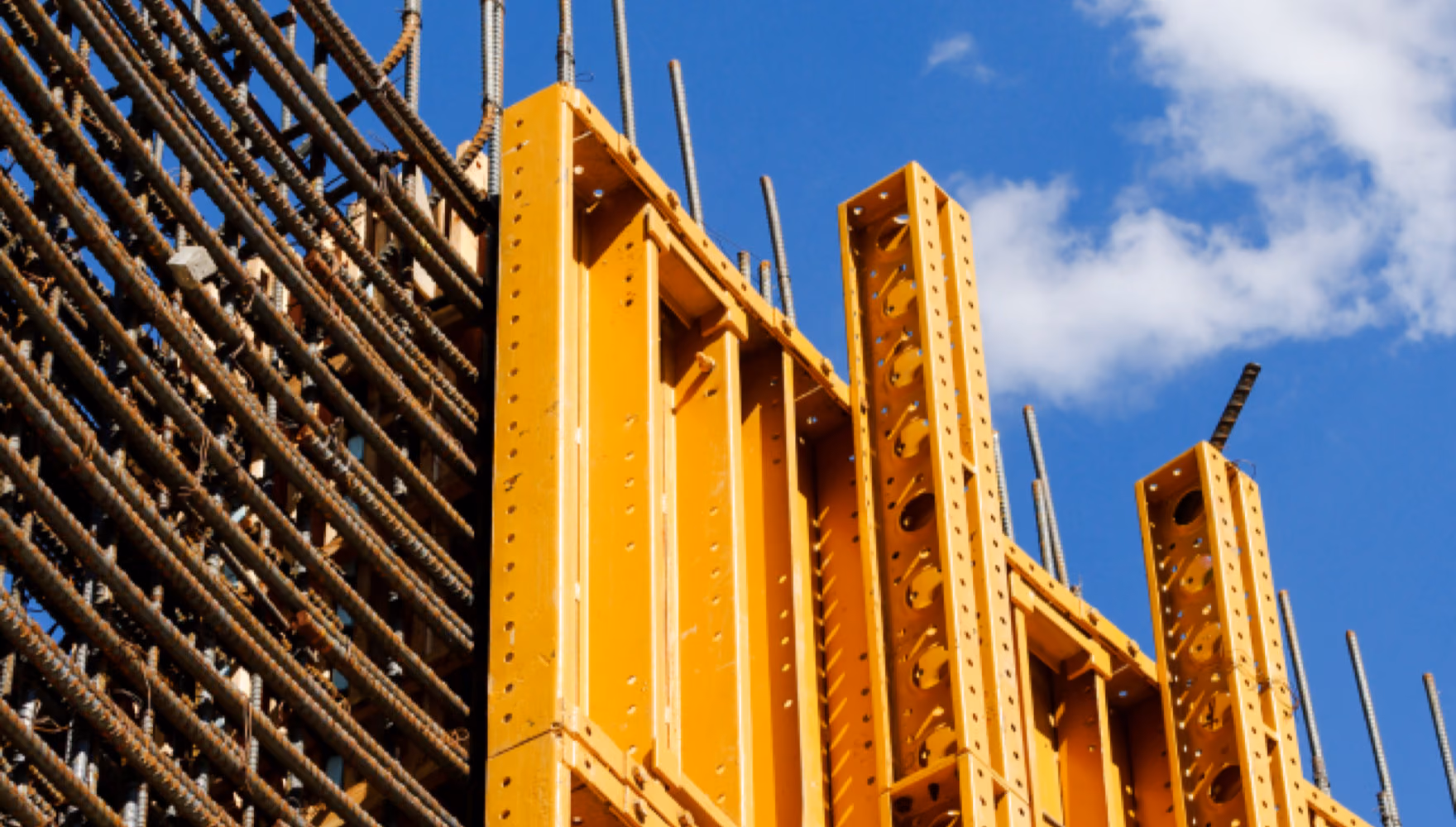 A dense cage of reinforcement bars butts against yellow steel wall formwork panels, rebar ends rising into a blue sky.