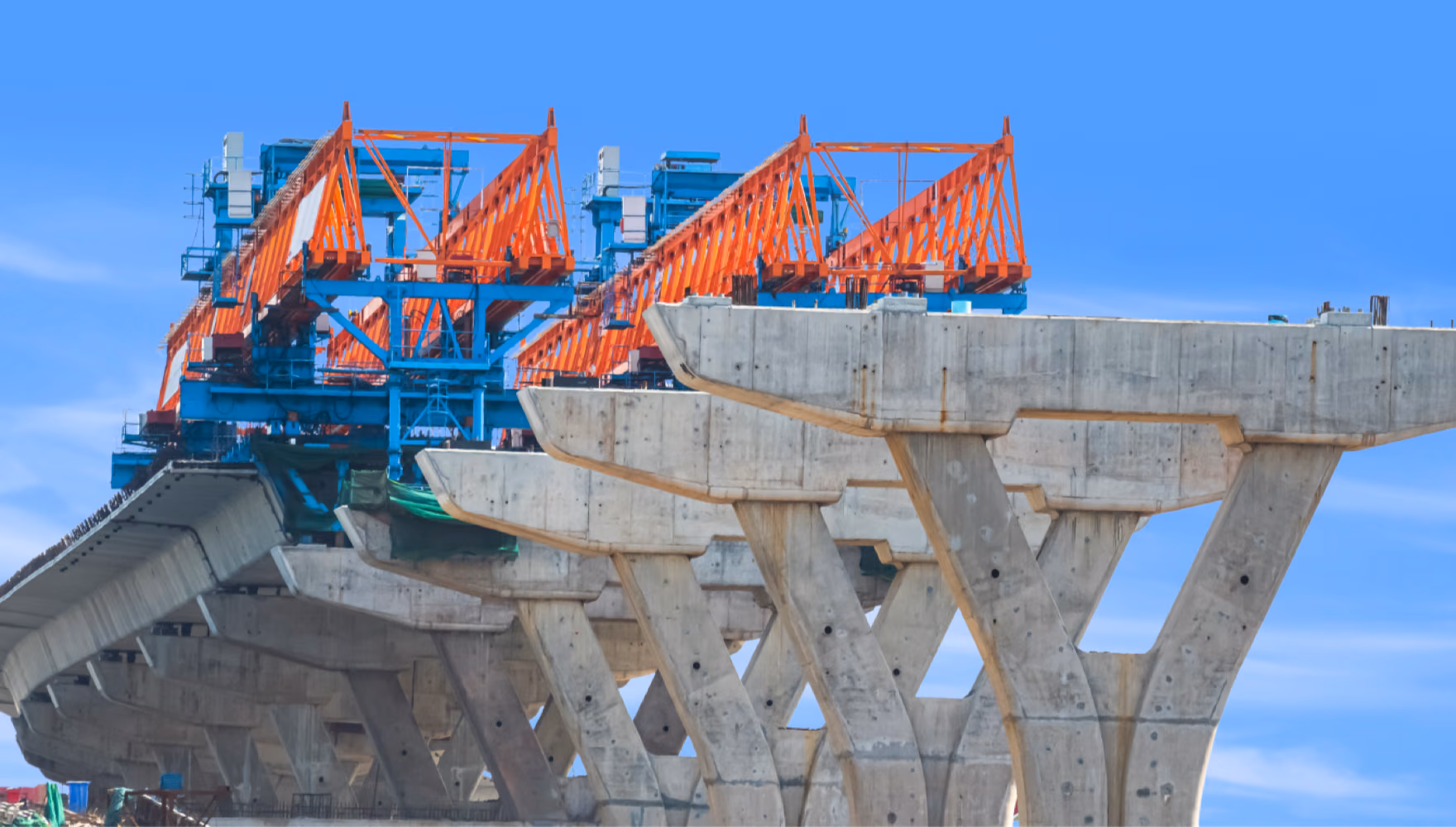 A bridge deck under construction, an orange launching gantry standing on a row of splayed concrete pier caps against a clear blue sky.