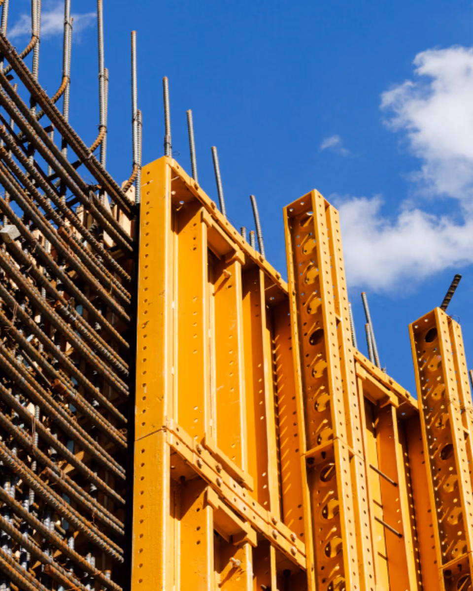 A dense cage of reinforcement bars butts against yellow steel wall formwork panels, rebar ends rising into a blue sky.