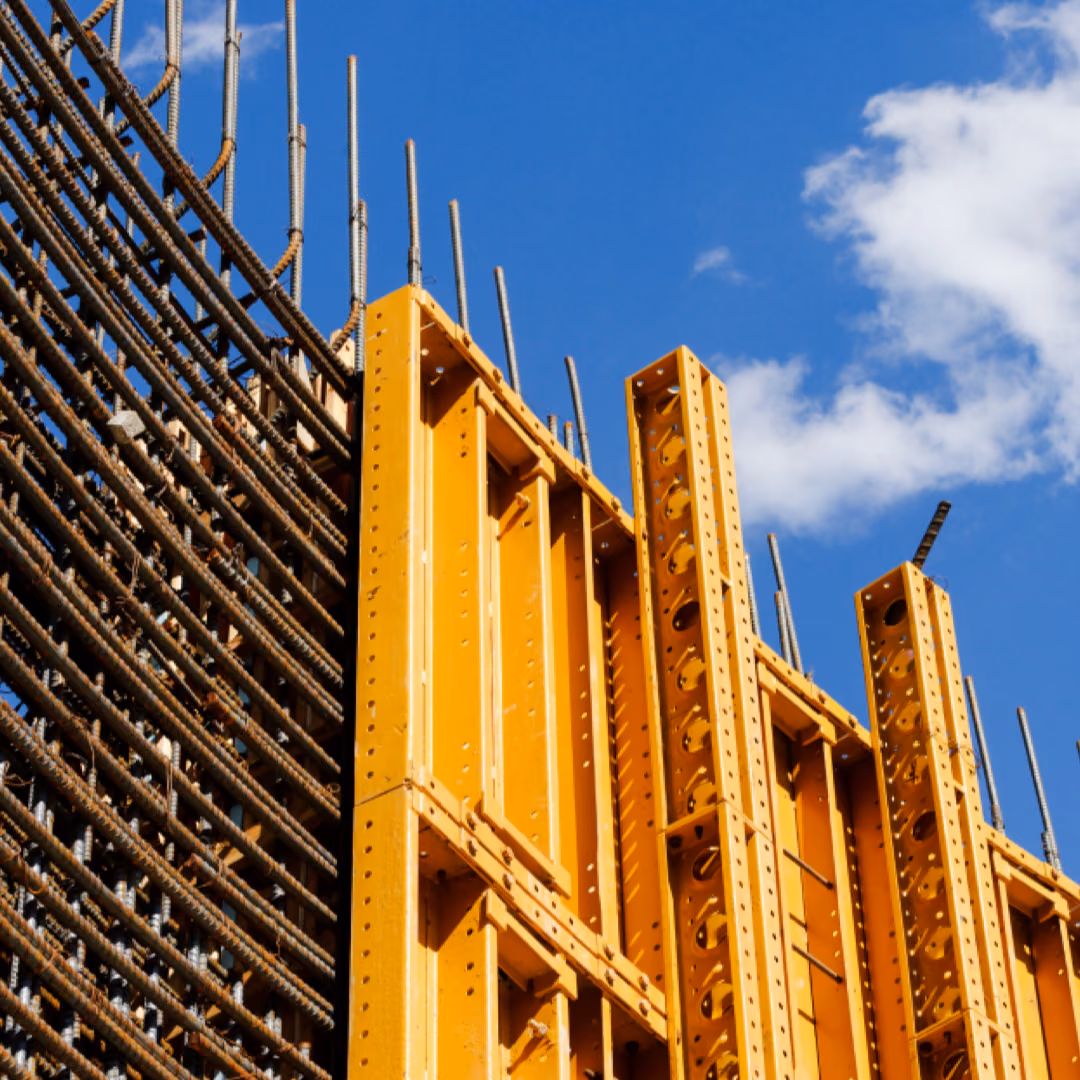A dense cage of reinforcement bars butts against yellow steel wall formwork panels, rebar ends rising into a blue sky.