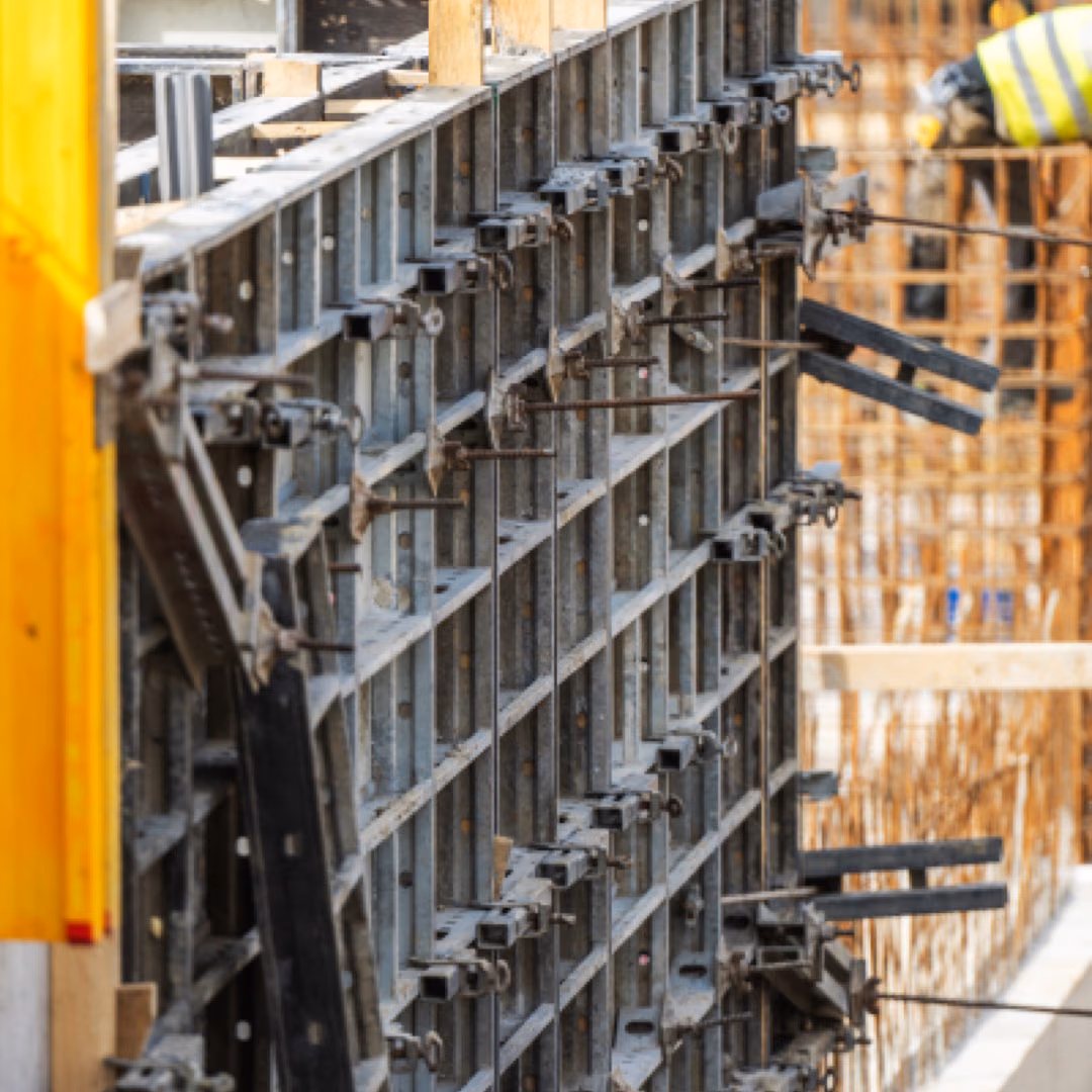 Steel wall formwork panels seen along their edge with tie rods and clamps in a row, a worker in a hi-vis vest out of focus behind.