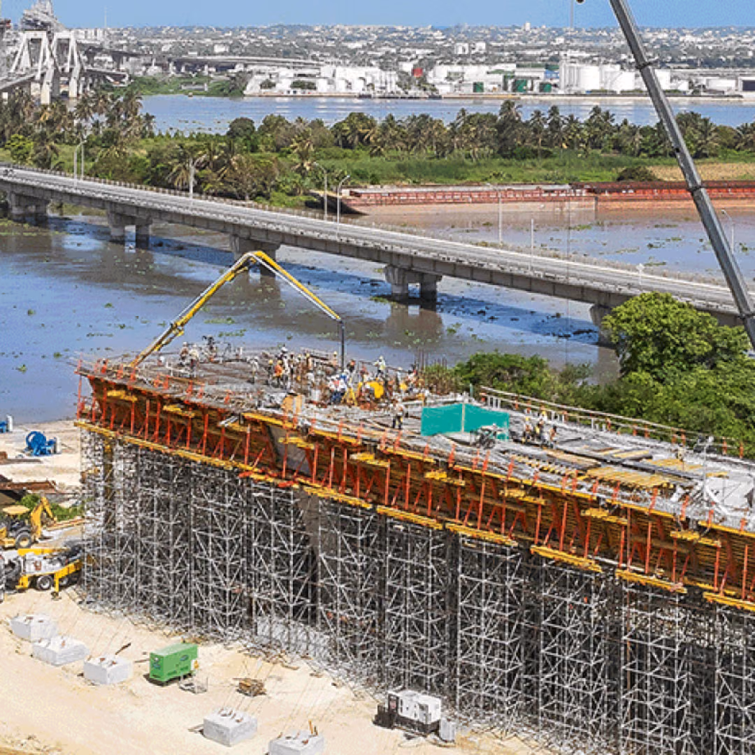An aerial view of a bridge site beside water, a long wall of red and yellow shoring props under the new deck, a concrete pump boom and a mobile crane at work, the existing bridge running behind it.