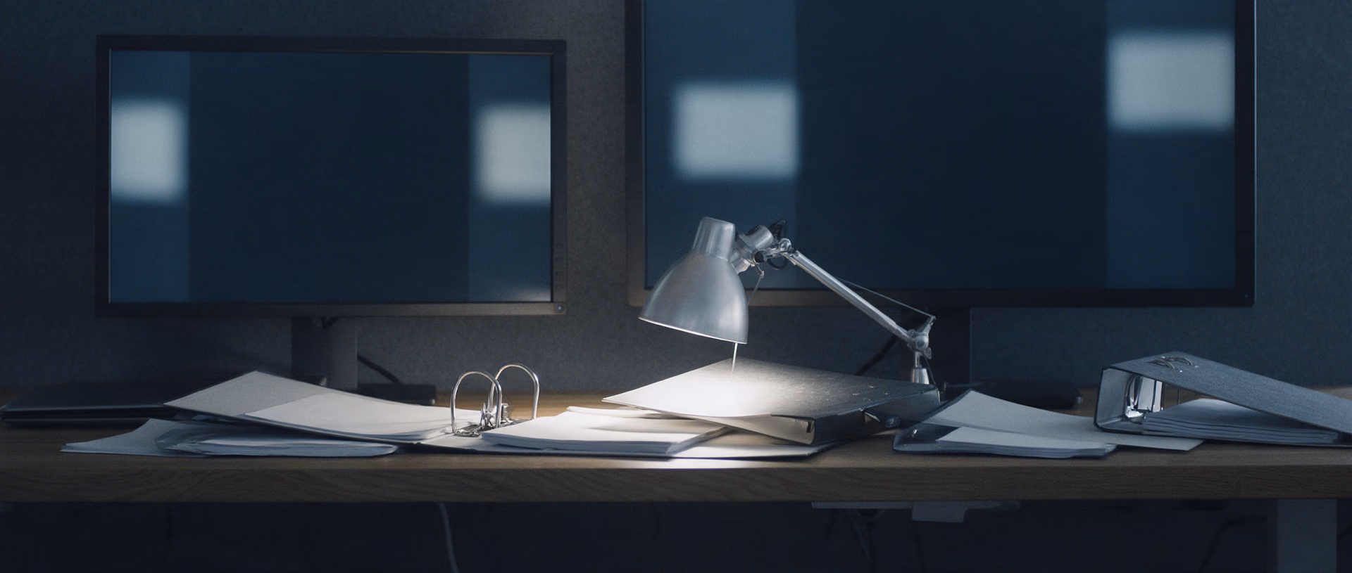 An empty office desk after hours with open ring binders and paper stacks under a desk lamp, two dark monitors standing against a grey partition.