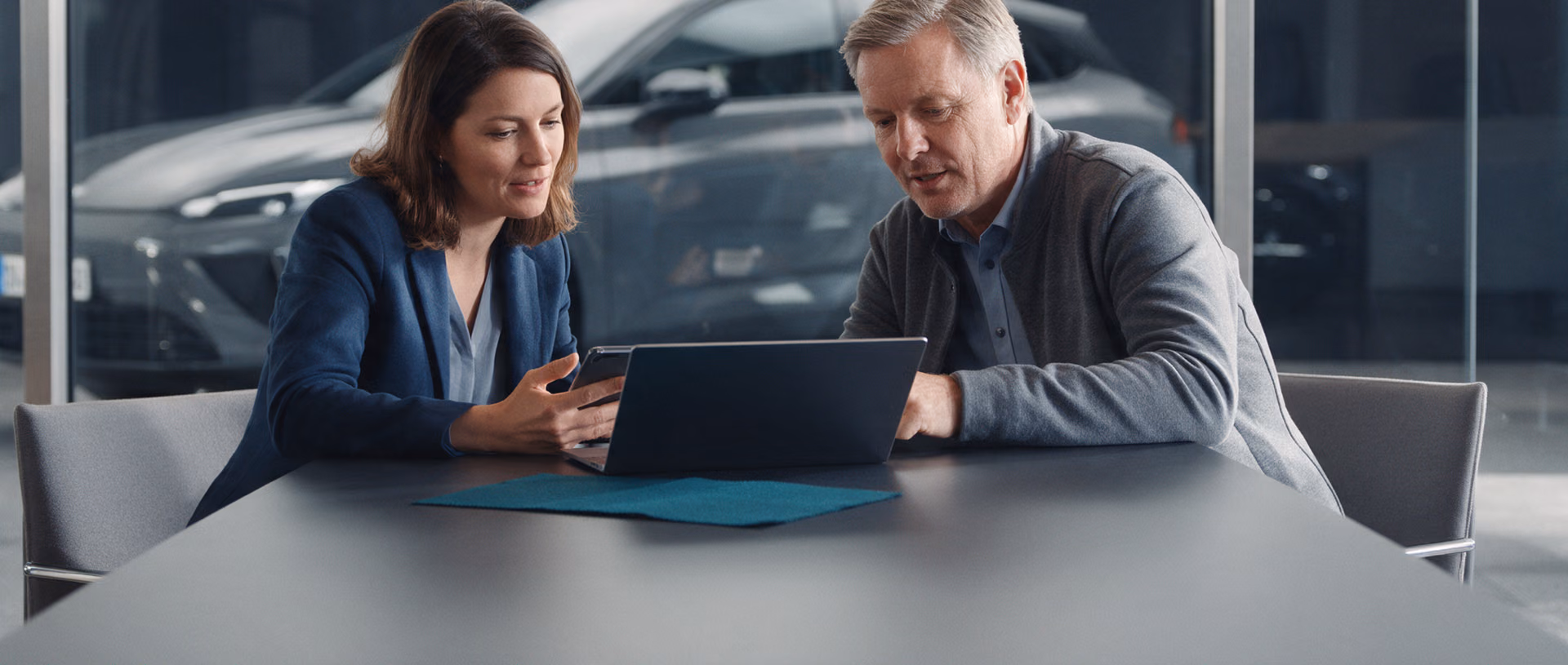 A saleswoman and a sales representative sit at a dark table in a car showroom looking at a laptop, a teal fabric swatch on the table and a grey car behind the glass.