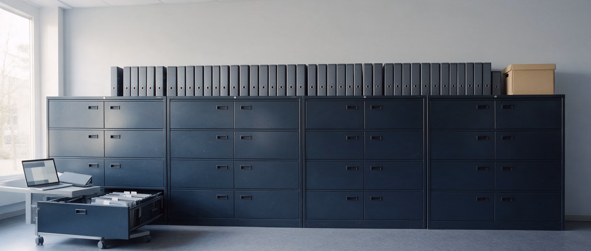 A row of dark steel filing cabinets with lever arch files and a cardboard archive box on top, a laptop on a low cart and one drawer pulled open.