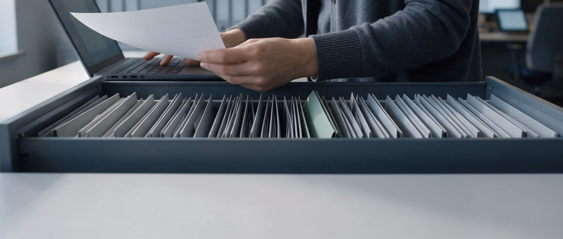 Hands hold a sheet of paper above a laptop keyboard in front of an open filing drawer of grey folders, one green folder standing among them.
