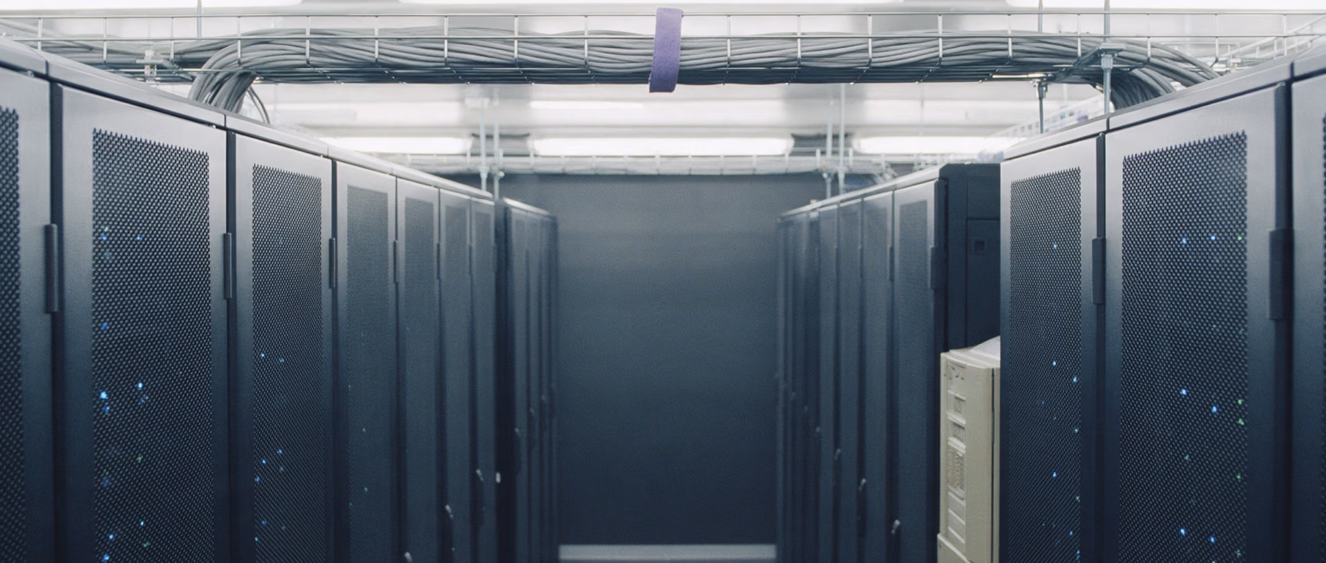 An aisle between two rows of dark server racks with closed perforated doors, a cable bundle on an overhead tray held by a violet strap.