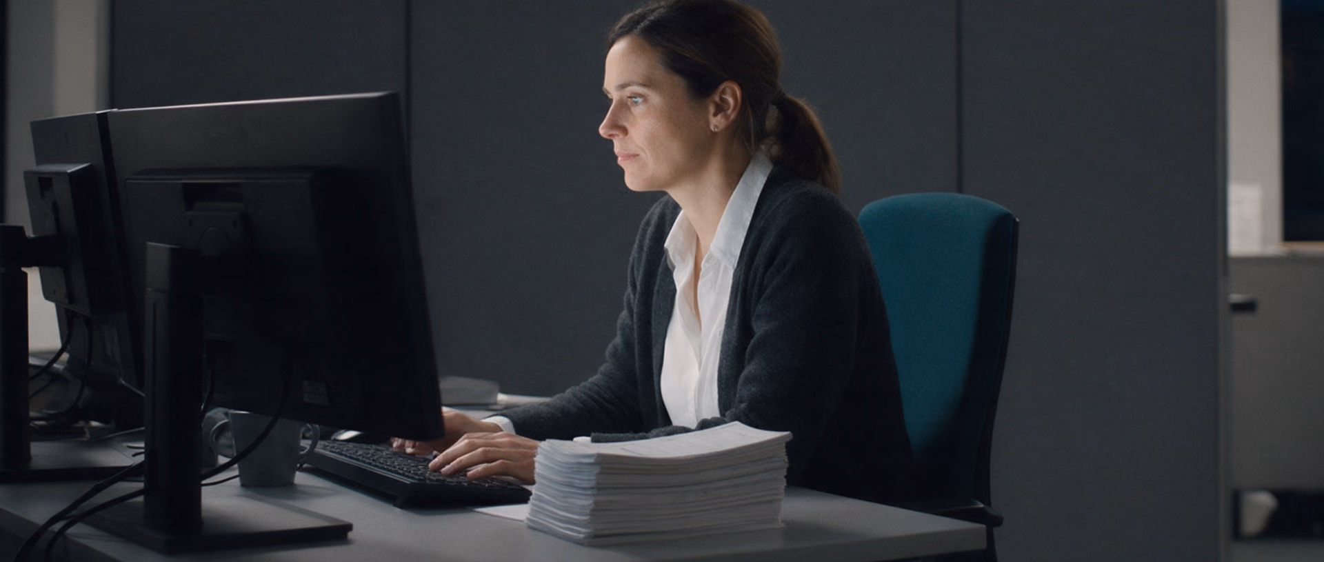 A woman in a white shirt and dark cardigan works at two monitors in an office after dark, a tall stack of paper beside the keyboard and a teal office chair behind her.