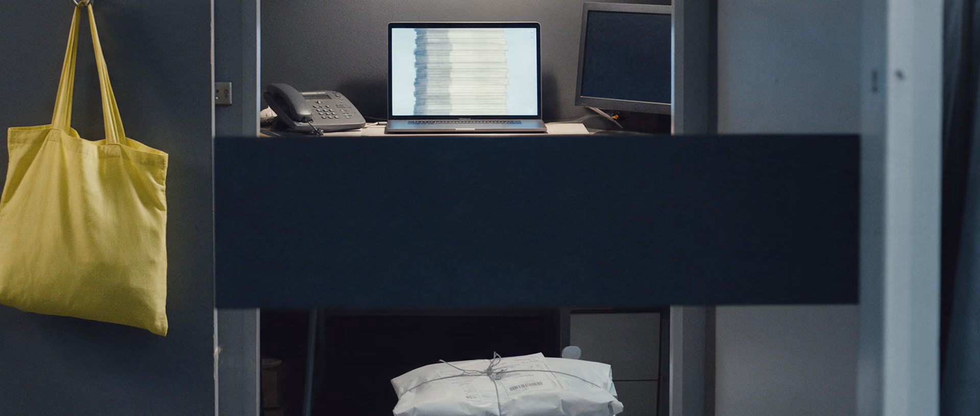 An empty back office behind a shop after closing, a glowing laptop and a dark monitor on the desk, parcels on the floor and a yellow tote bag on a wall hook.