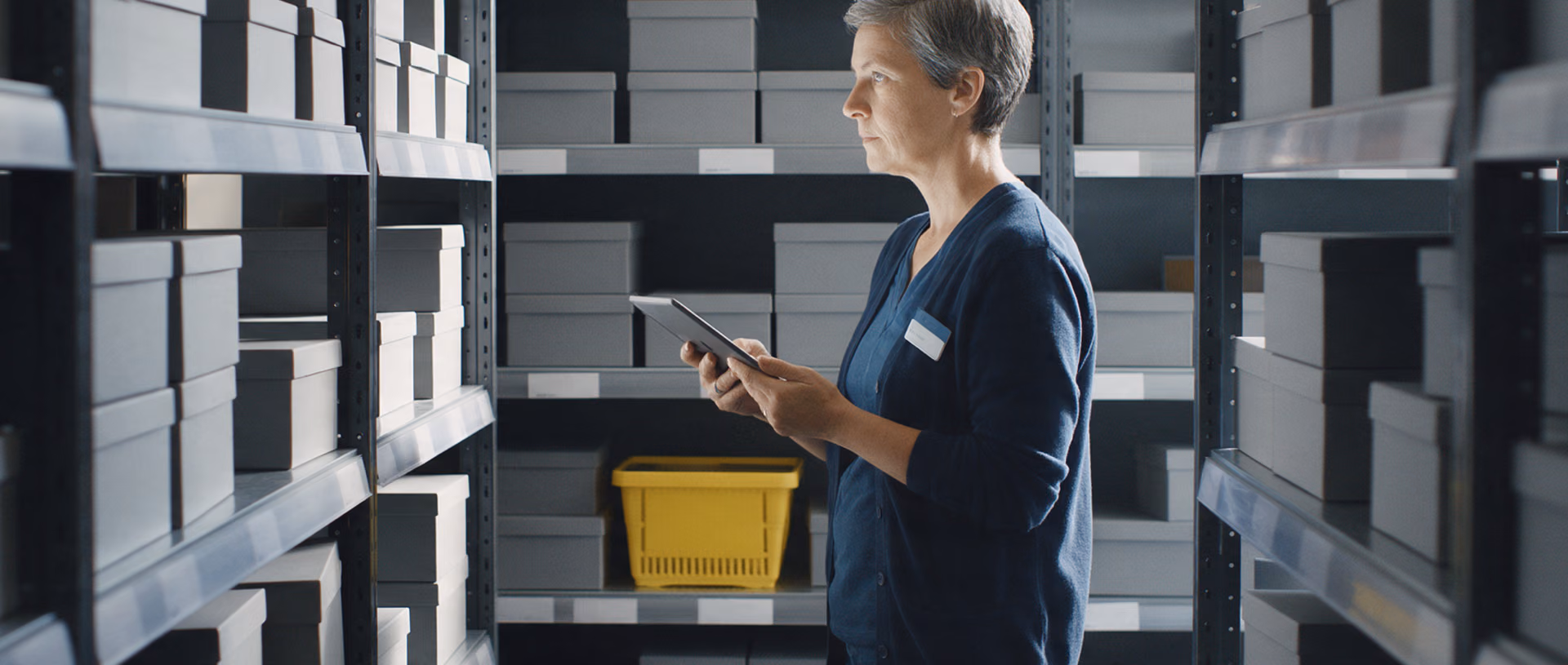 A store employee stands between grey stockroom shelving units holding a tablet and looks at the stacked boxes, a yellow shopping basket on the lower shelf beside her.