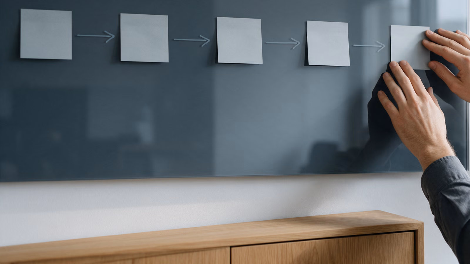 Hands placing a blank sticky note at the end of a chain of notes joined by arrows on a dark glass board.