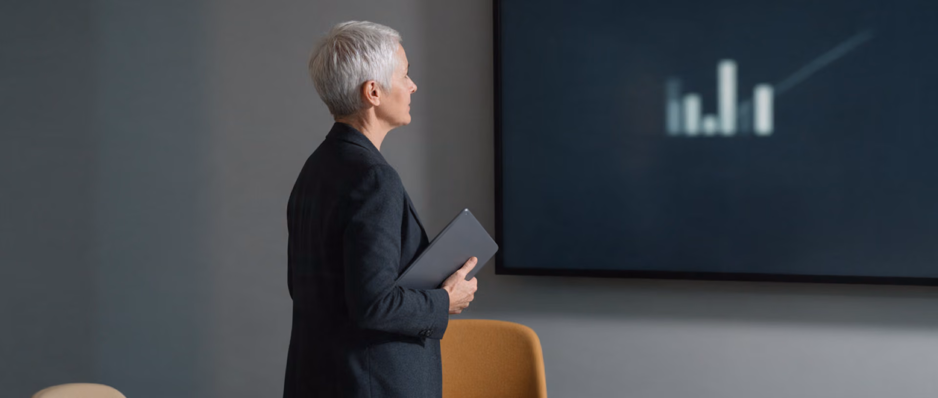 A woman with a tablet stands facing a wall screen showing blurred bars of unequal height in a quiet meeting room.