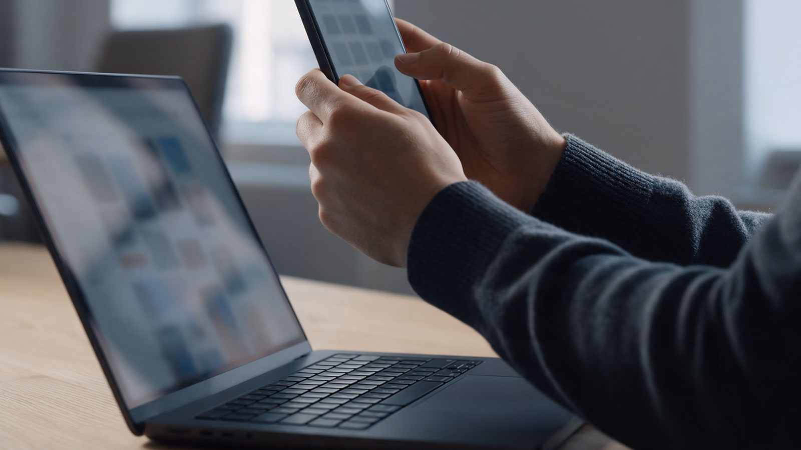 Hands holding a smartphone above a laptop on a wooden desk.