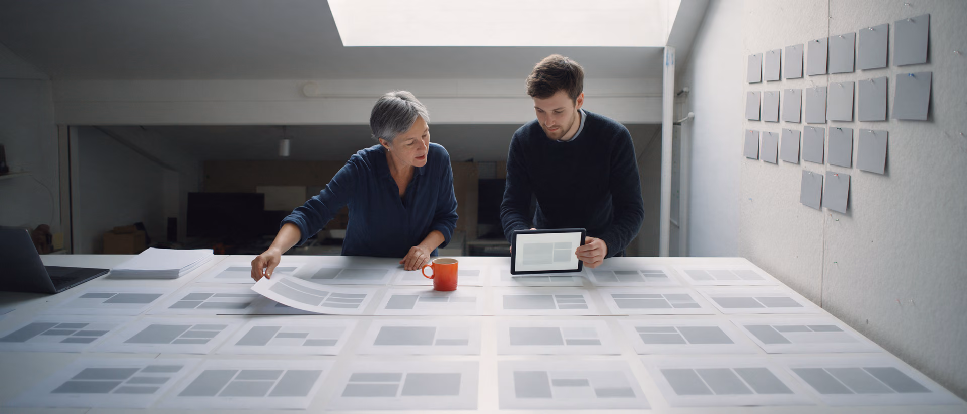Two colleagues lean over a long white studio table covered in rows of blank layout sheets, one holding a tablet, an orange mug between them and a wall of pinned blank cards to the right.