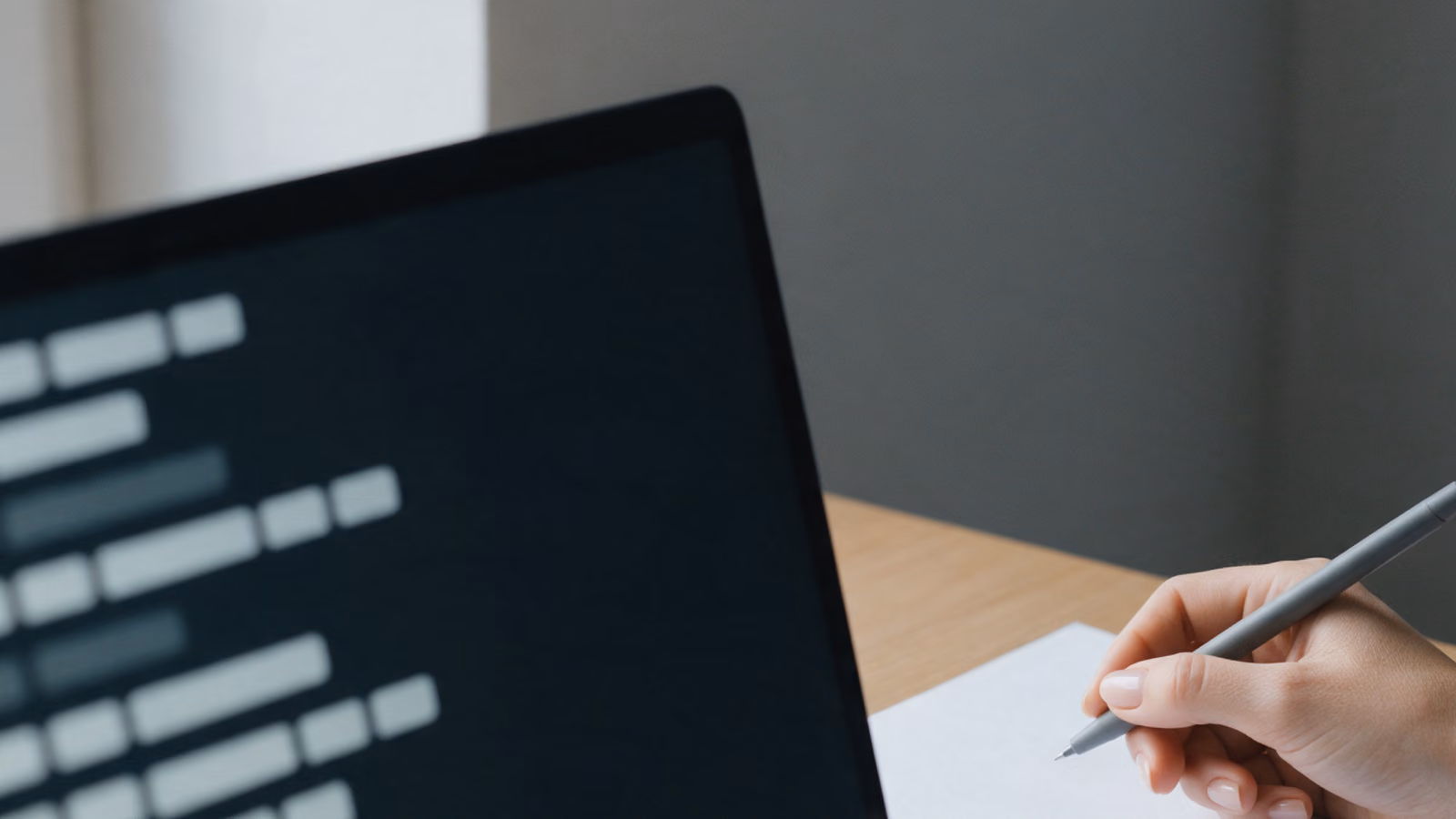 Close view of a laptop showing blurred list rows beside a hand holding a pen over a blank sheet of paper on an oak desk.