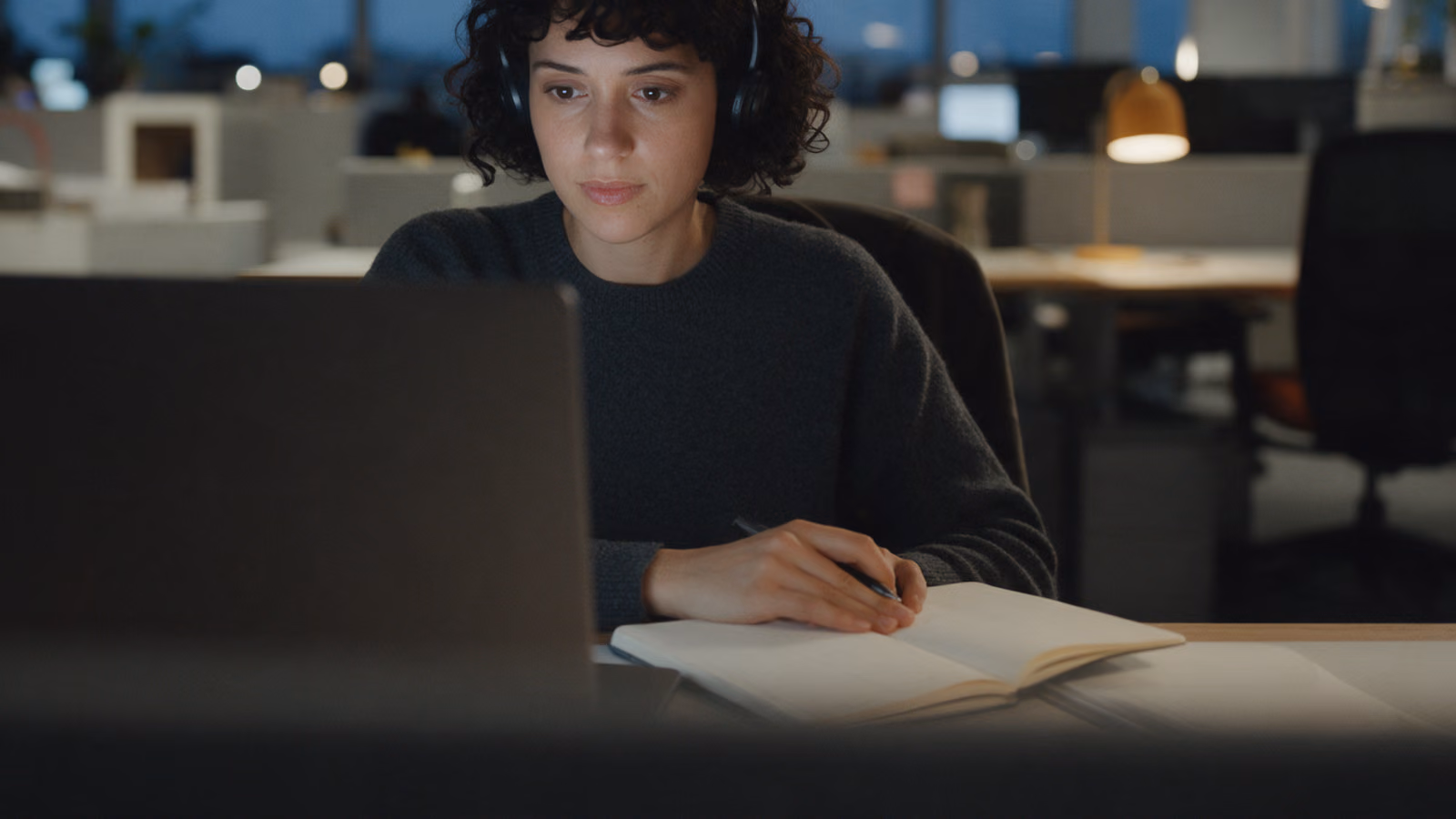 A woman with headphones works alone at her laptop in a quiet office at dusk, a pen over an open notebook and a small desk lamp beside her.