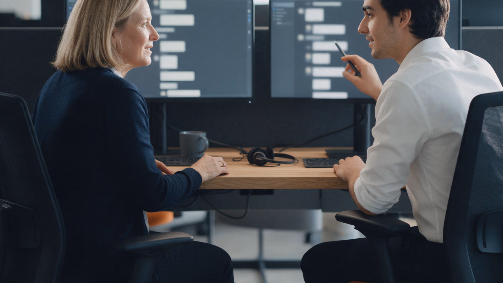 Two colleagues at neighbouring desks turn towards each other to talk, their monitors showing blurred stacks of message-like blocks.