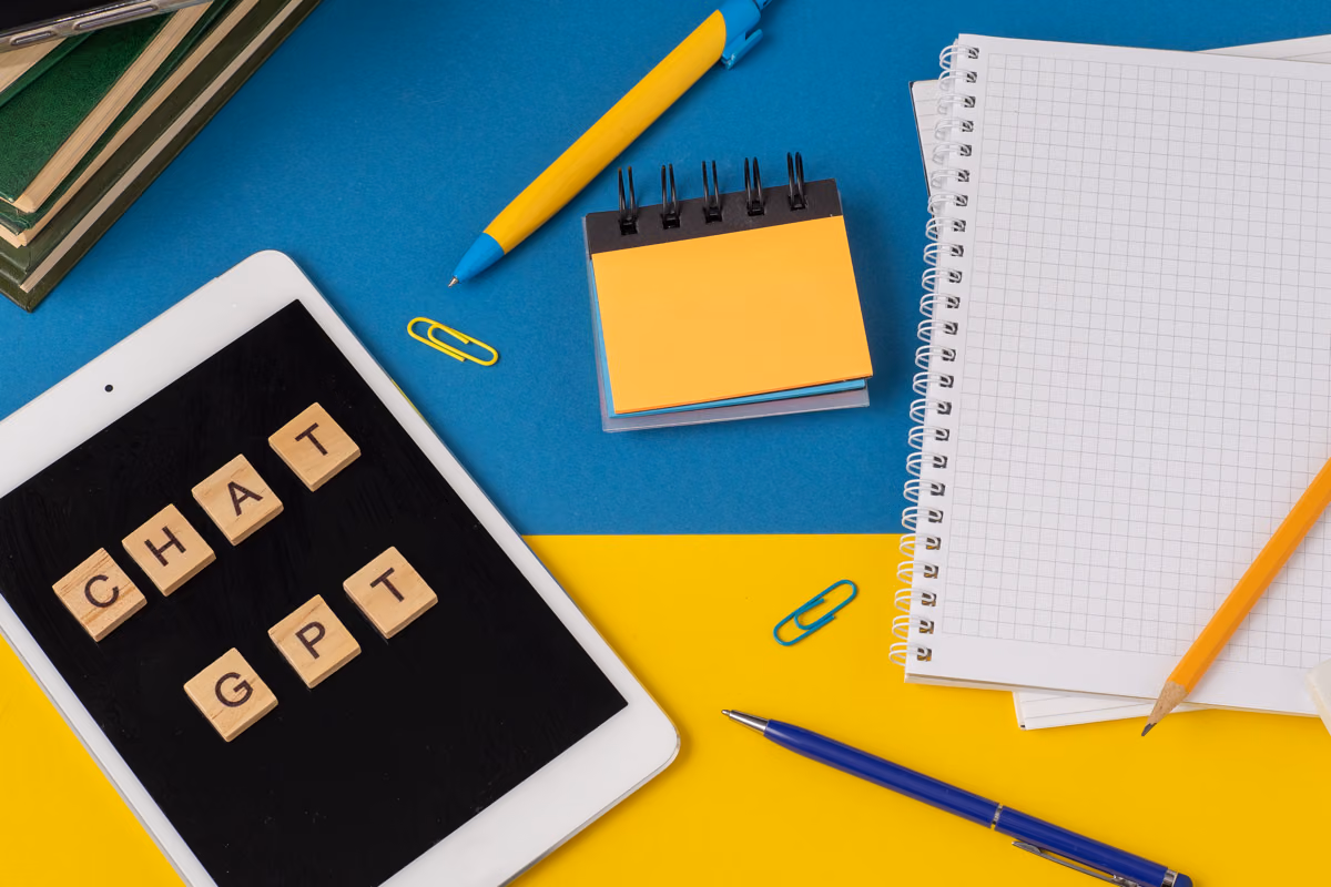 Wooden letter tiles spelling “chat GPT” on a tablet beside notebooks, pens and paper clips.