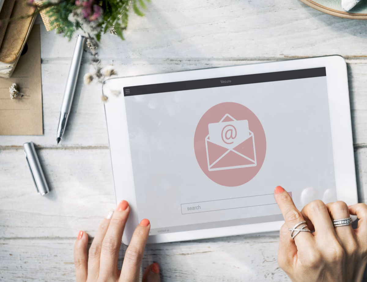 Two hands holding a tablet that shows an email icon, on a white wooden desk.