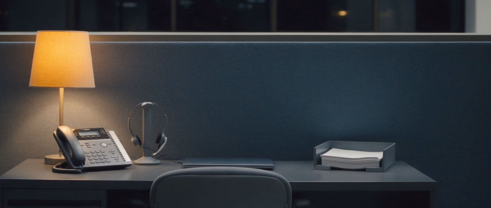 An unoccupied service desk in an office after dark, a desk phone, a headset on its stand, a closed laptop and a tray of sheets under a lit desk lamp.