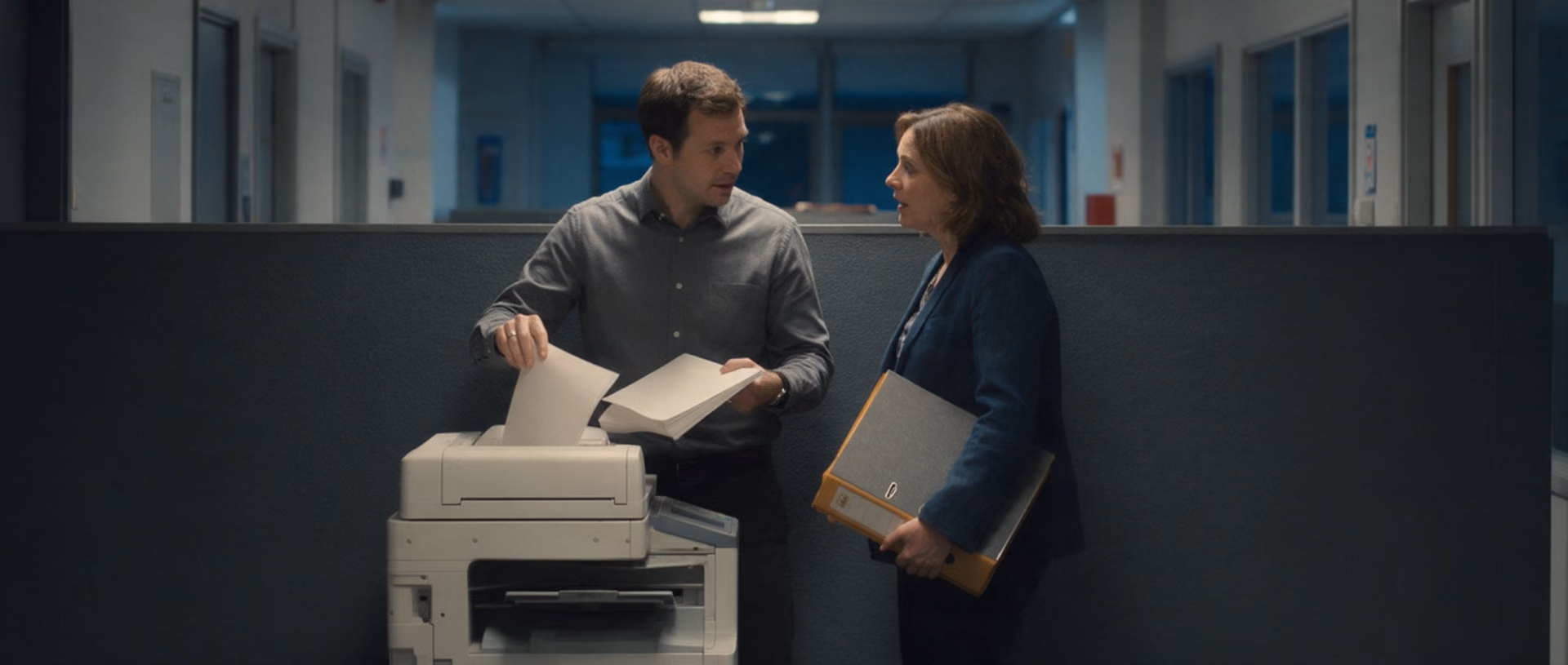 Two colleagues talk at a printer station in an office corridor after dark, one lifting a stack of sheets from the tray, the other holding a binder against her hip.