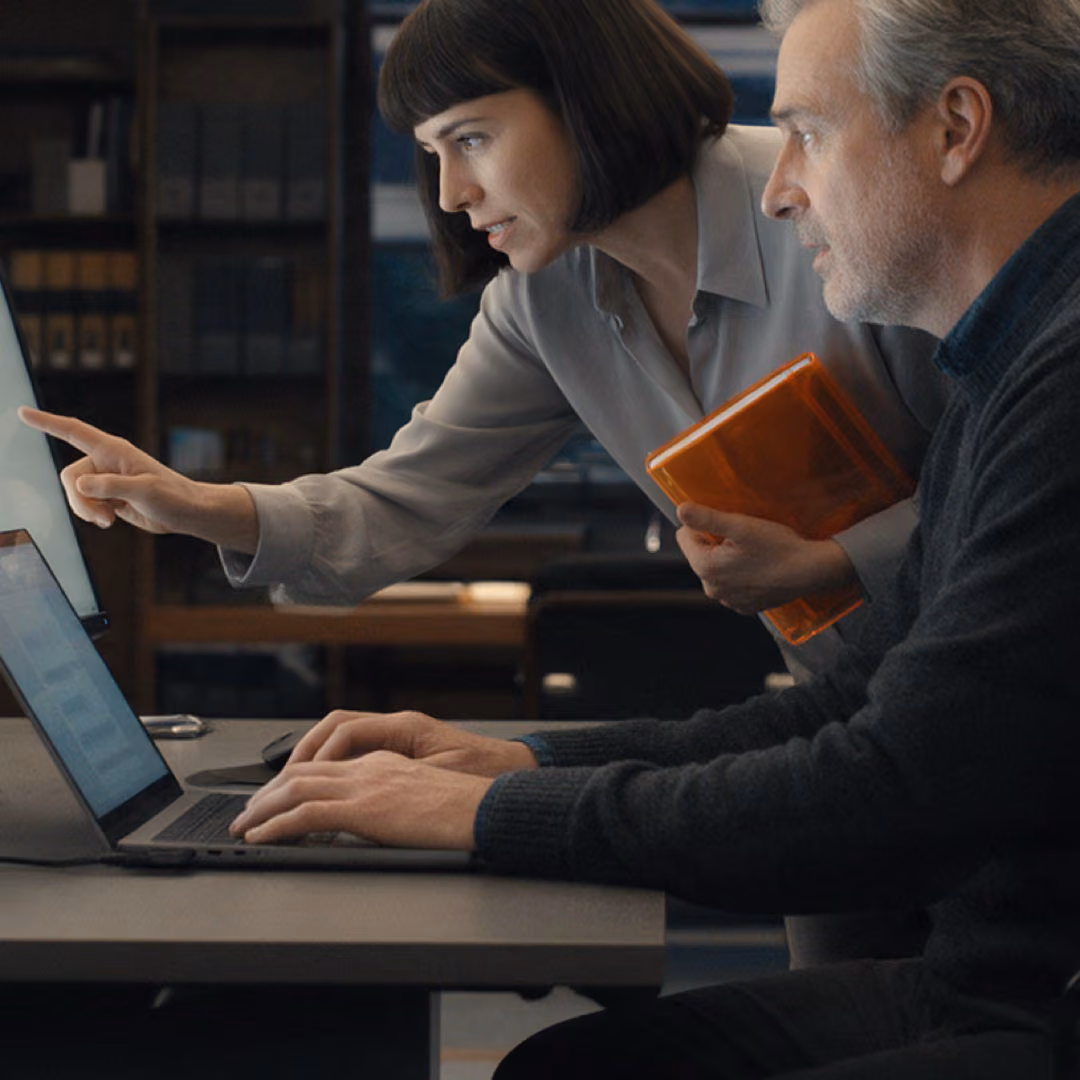A woman standing beside a seated colleague points at his monitor while he types on a laptop, an orange notebook under her arm.