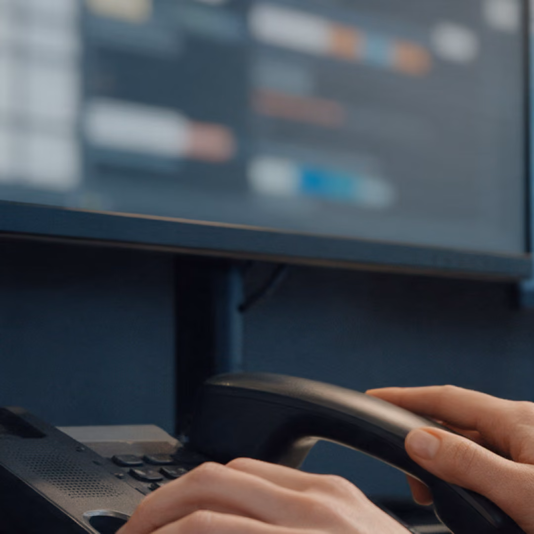 Two hands lift a desk phone handset beneath a monitor showing blurred rows of blocks.