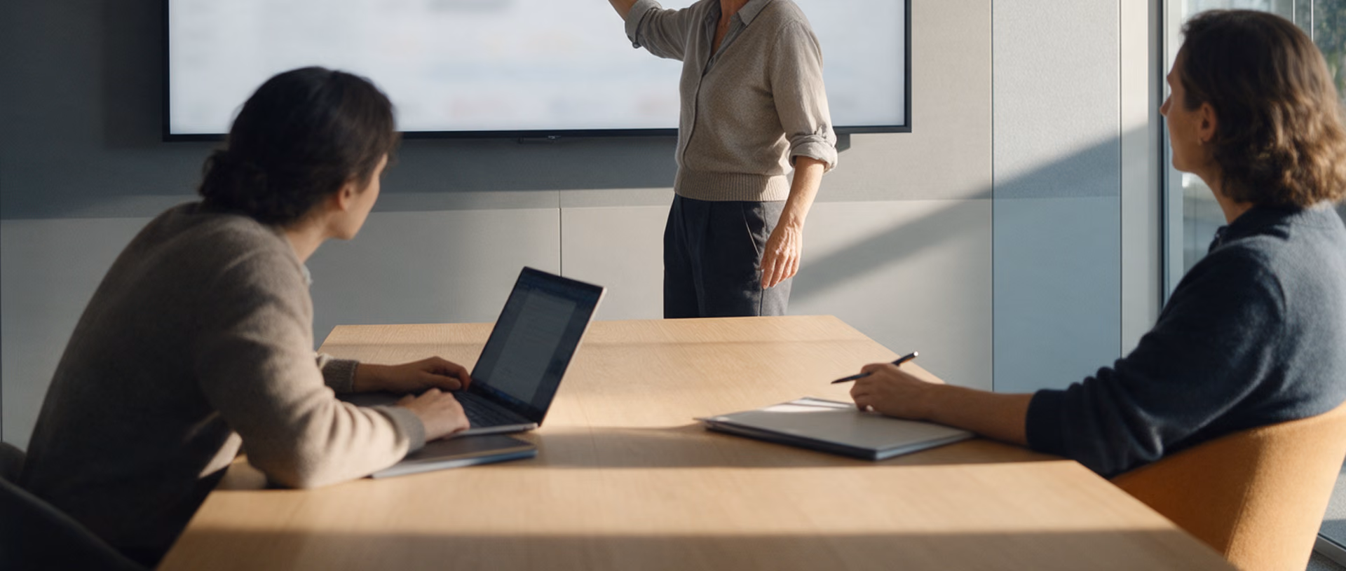A woman stands beside a large wall display in a glass-walled meeting room, one hand raised towards it, talking to two seated colleagues with an open laptop.