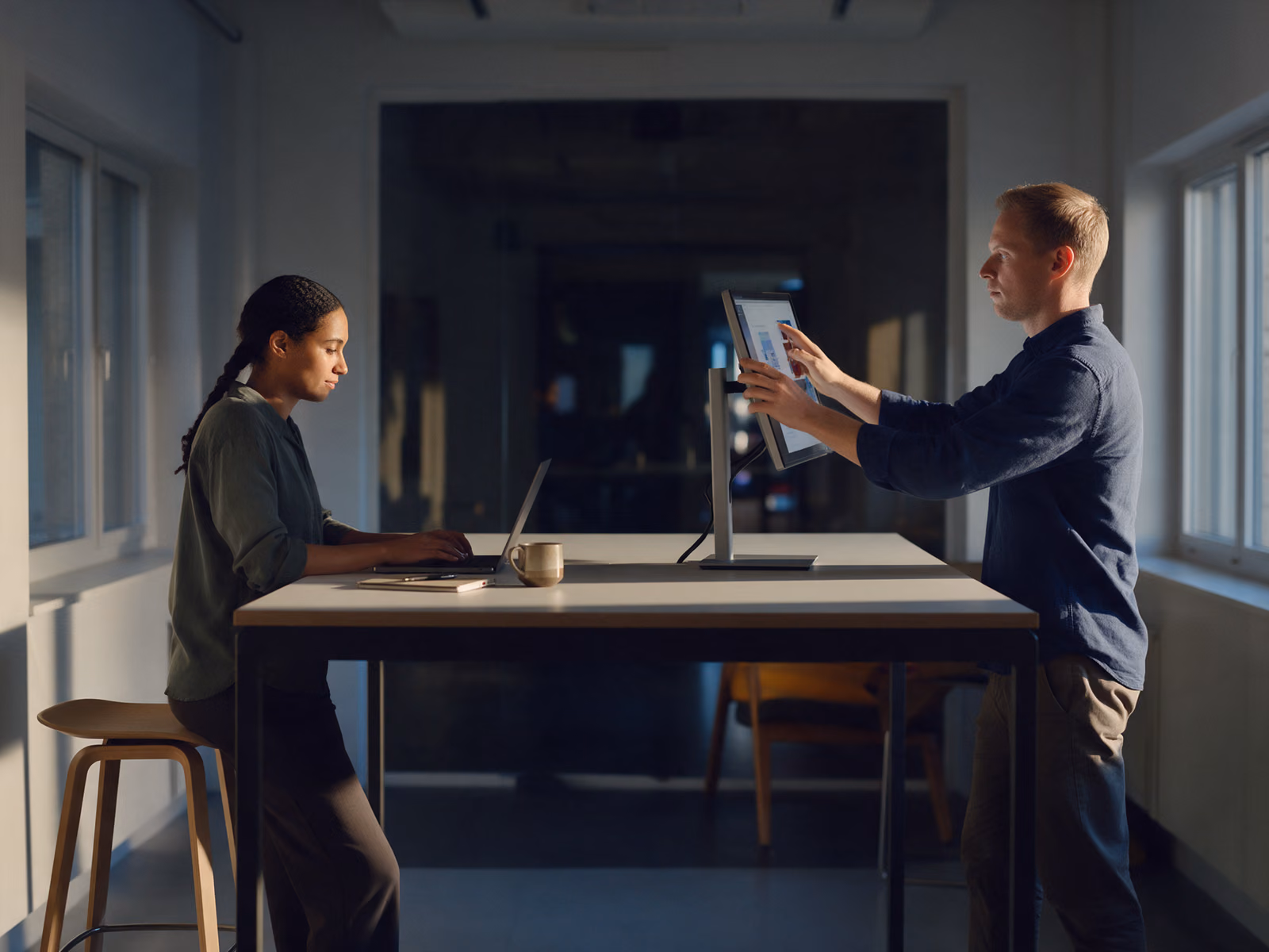 Two people at a standing desk, one typing on a laptop, the other turning a monitor on a stand towards her.