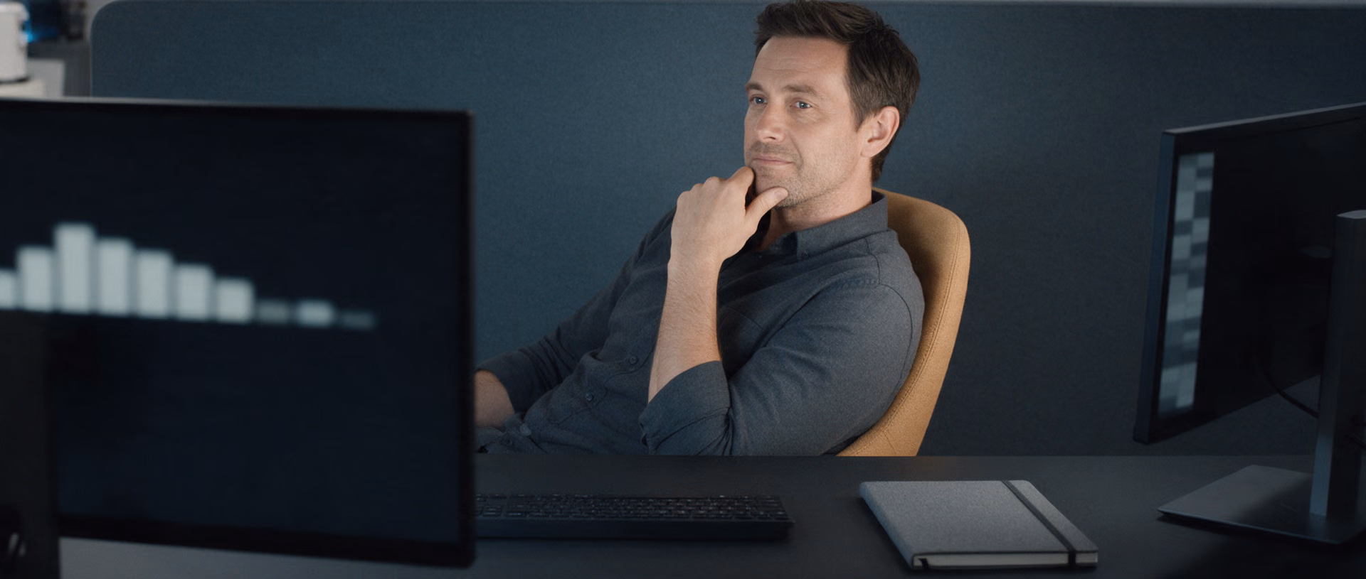 A man leans back in an office chair after dark, hand on his chin, looking between a large monitor with a row of blurred bars and a smaller monitor with a column of blocks.