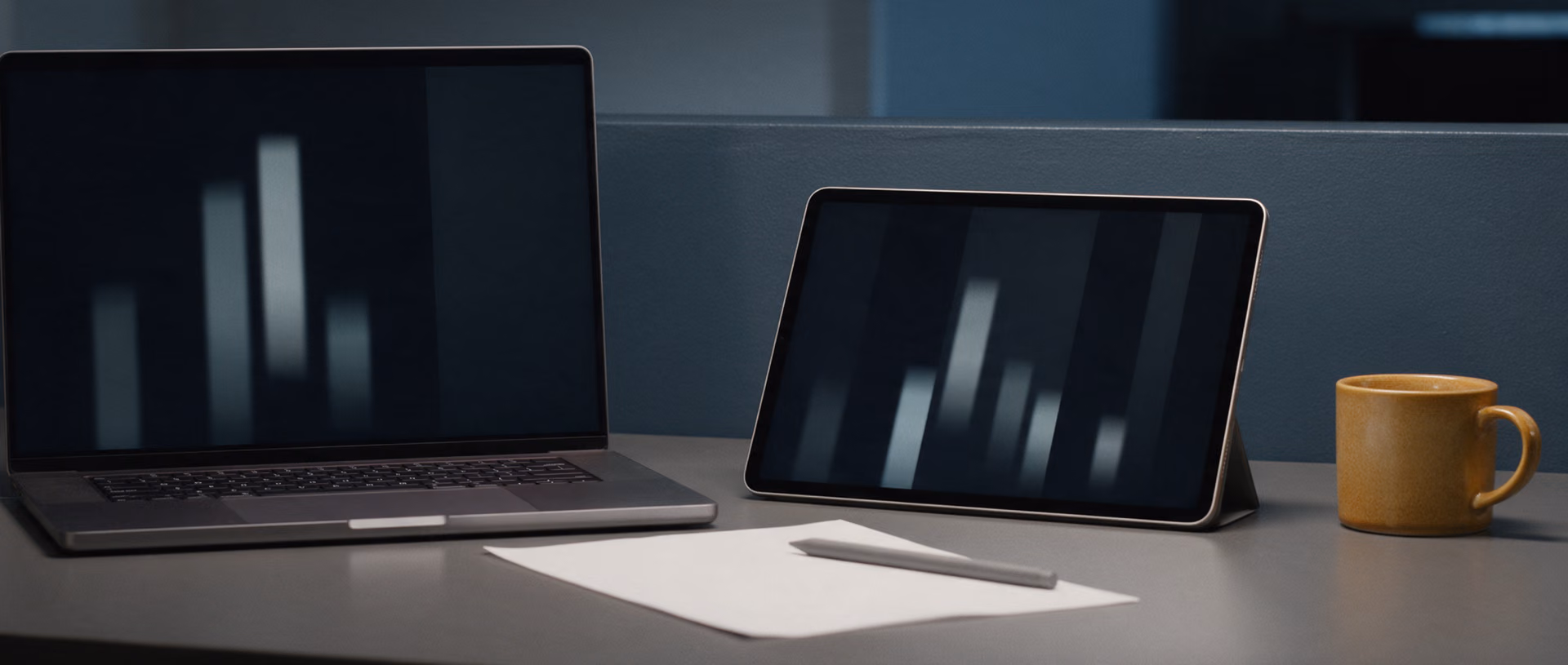 An open laptop and a tablet stand side by side on a dark desk, each screen showing a different set of blurred bars, a blank sheet and pen between them and a mug at the edge.