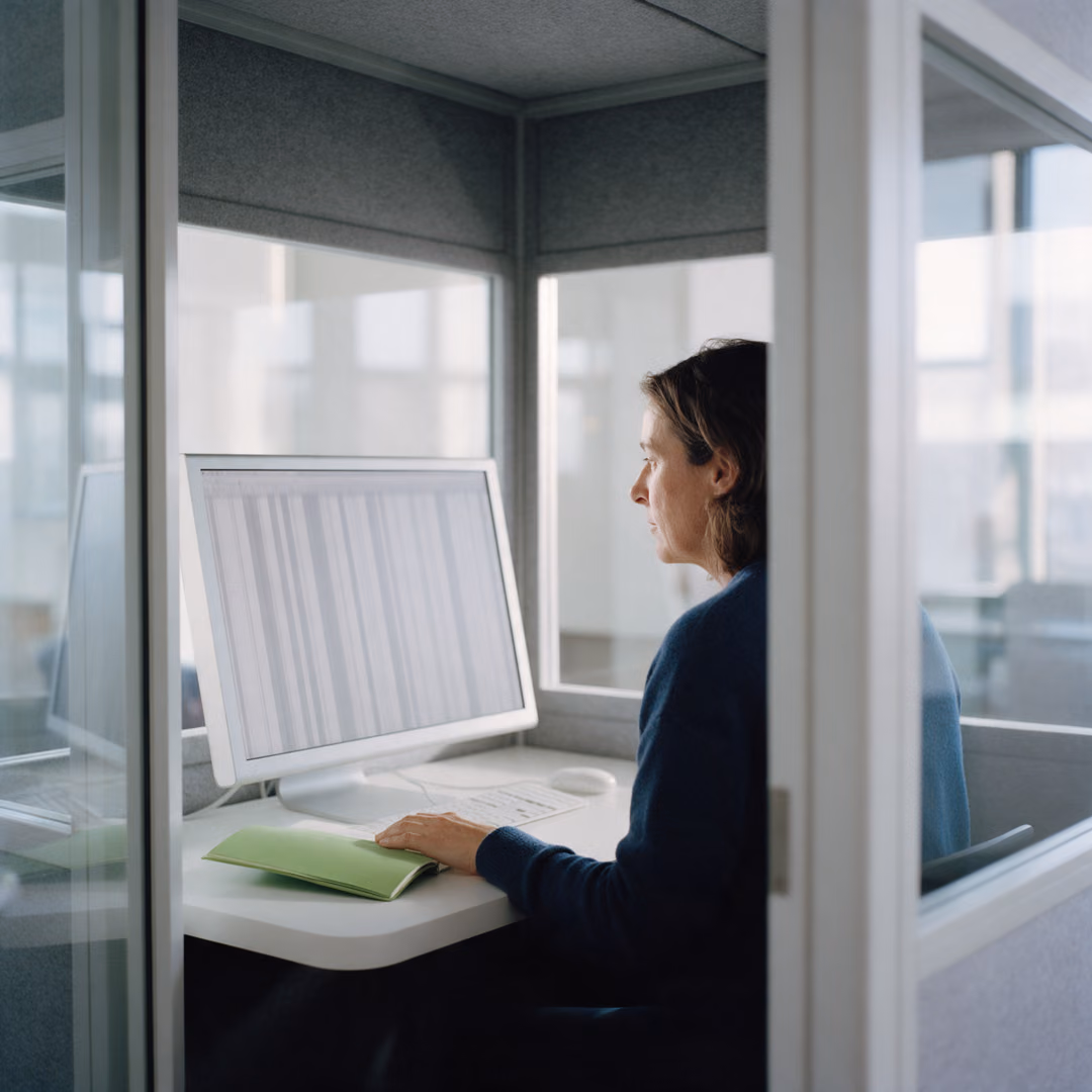 A woman sits in a glass focus booth reading columns of blurred rows on a large monitor, a green notebook beside the keyboard.
