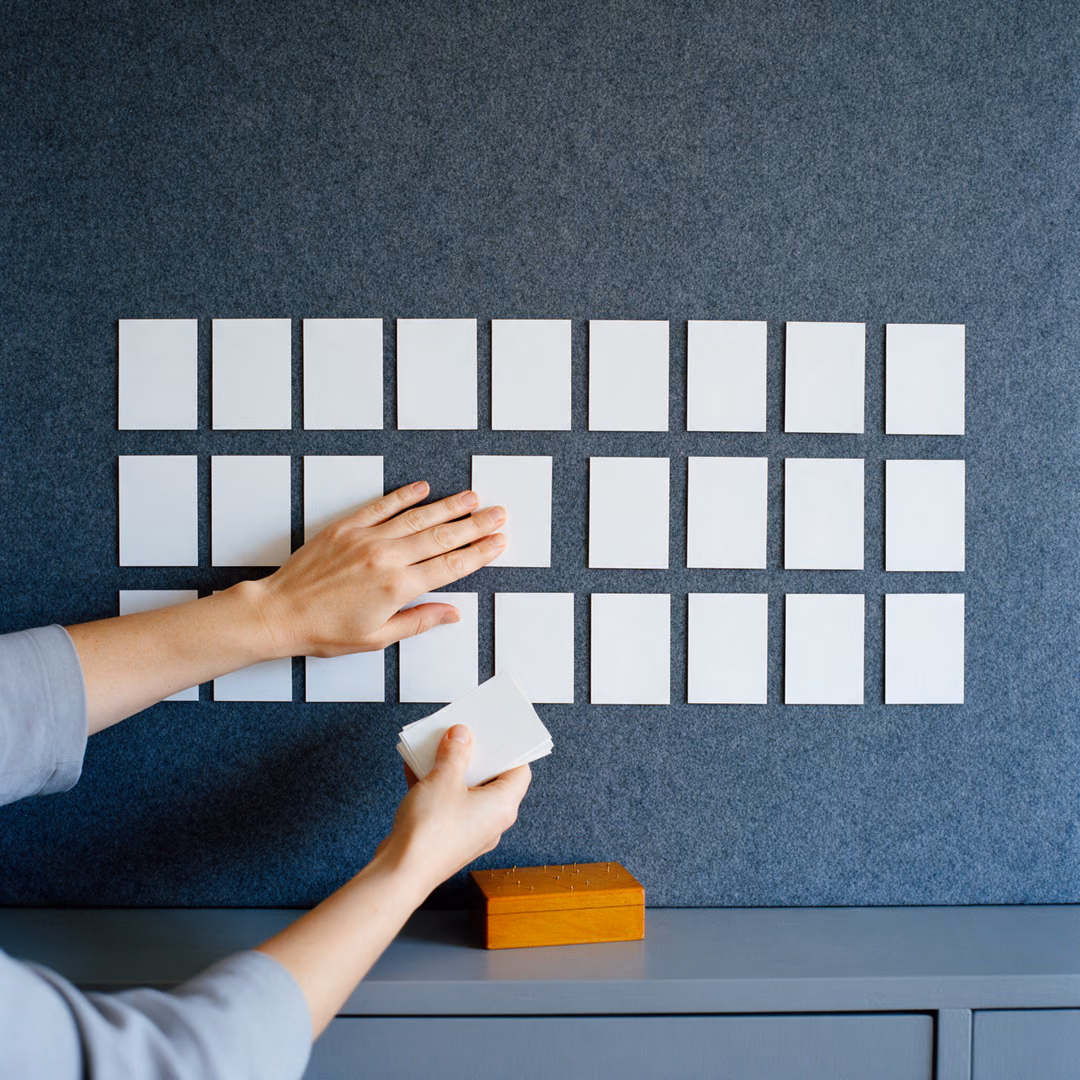Two hands press a blank white card into one of three rows of cards on a grey felt wall, a small wooden box of pins on the sideboard below.