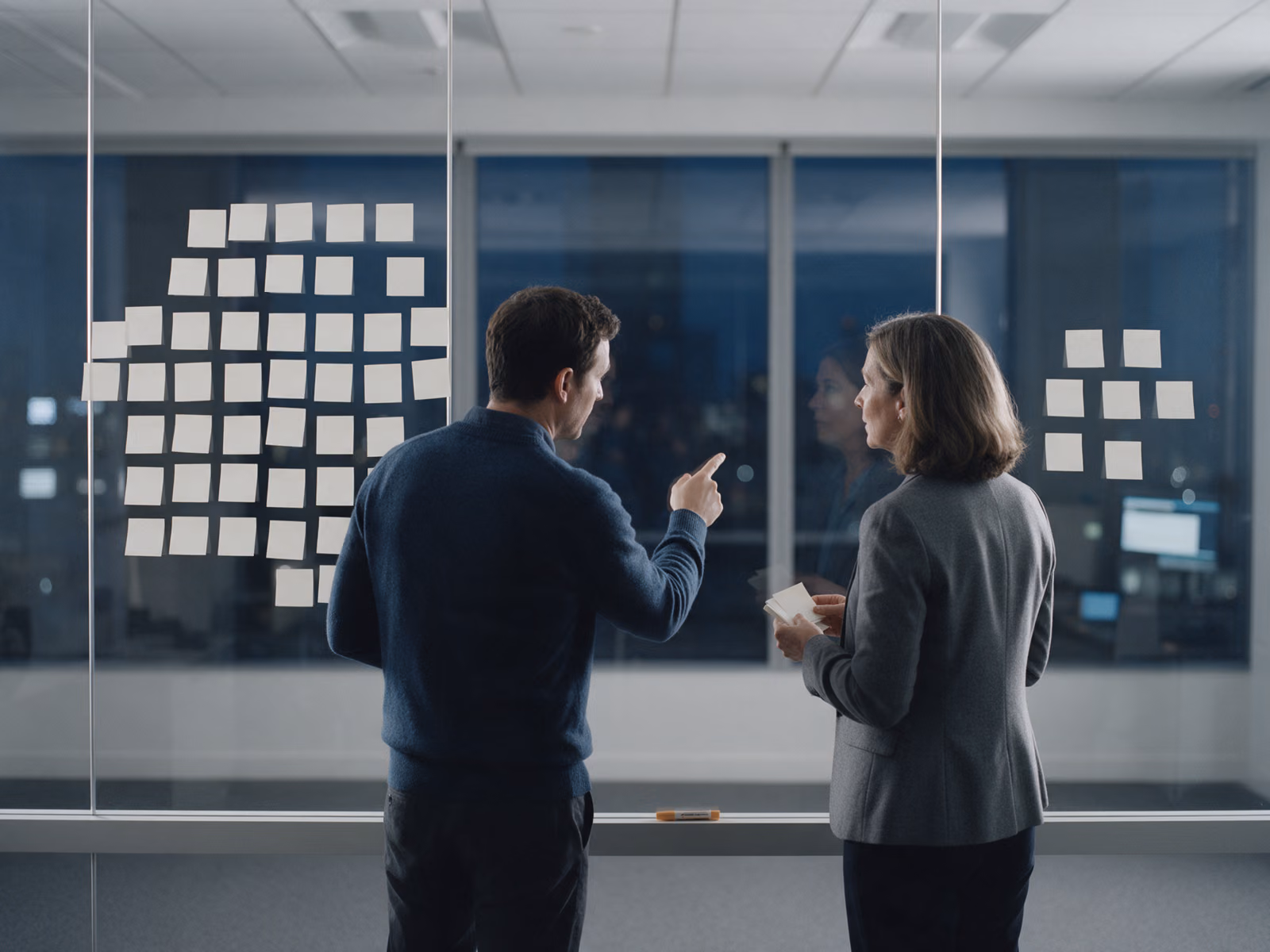 Two people stand at a glass wall after dark, blank notes arranged in two clusters with an empty stretch of glass between them, one pointing at the gap.